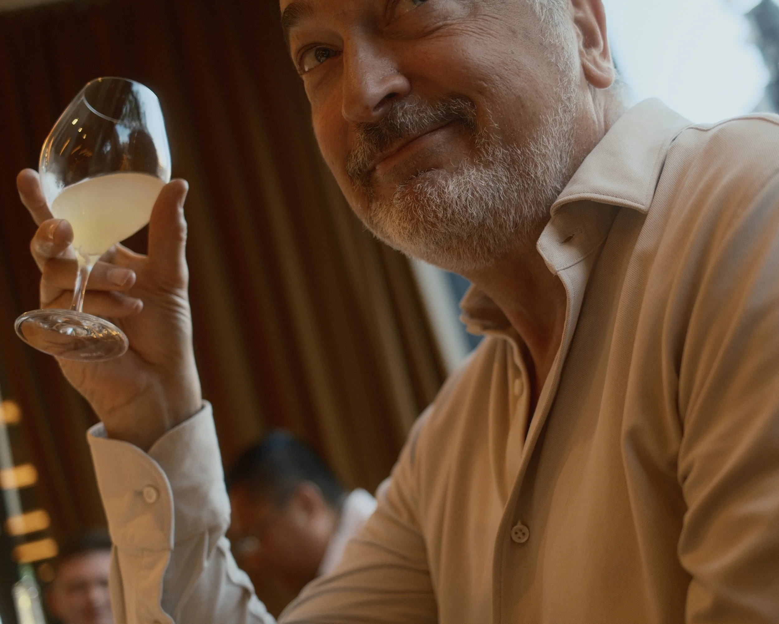 Close-up of a man with gray hair and beard holding a glass of white wine, smiling with eyes slightly squinted.