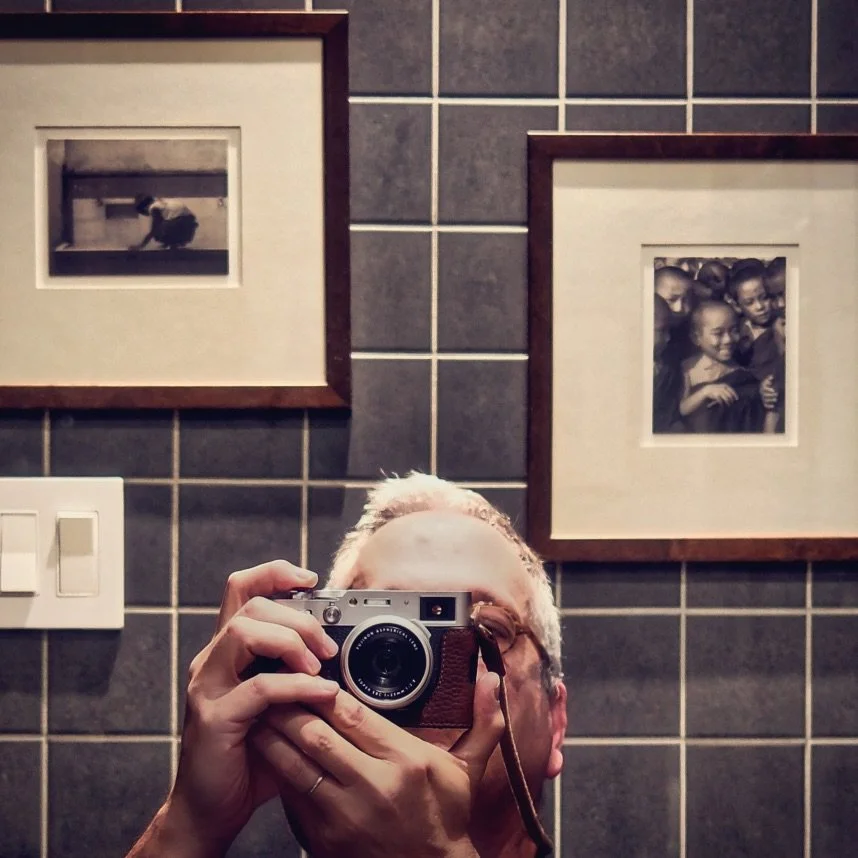 Person taking a photo with a vintage camera in front of a mirror, with framed black-and-white photographs on a tiled wall behind him.