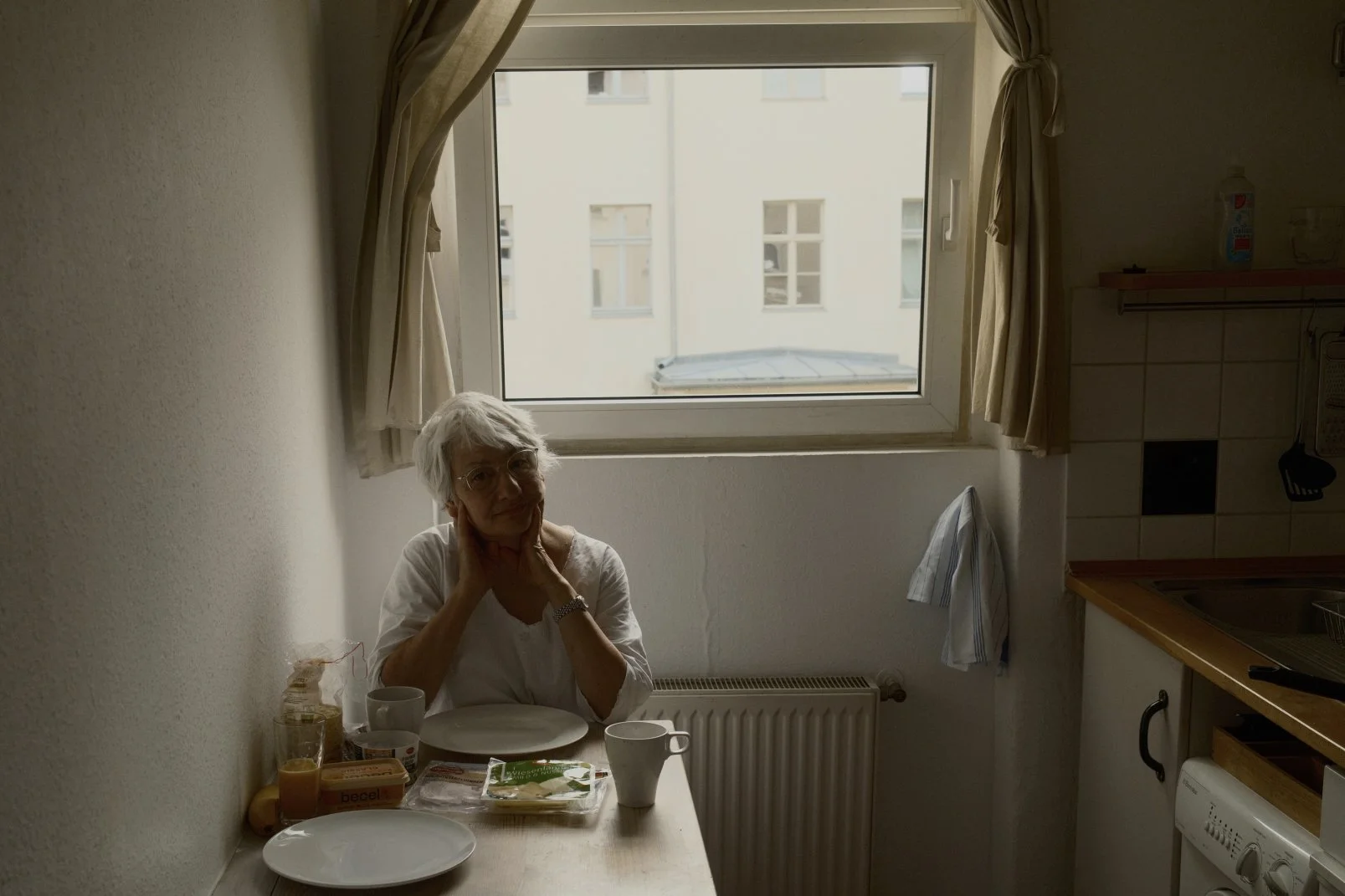 An elderly woman with glasses and gray hair sitting at a kitchen table near a window, with breakfast items including orange juice, bread, and cups in front of her.