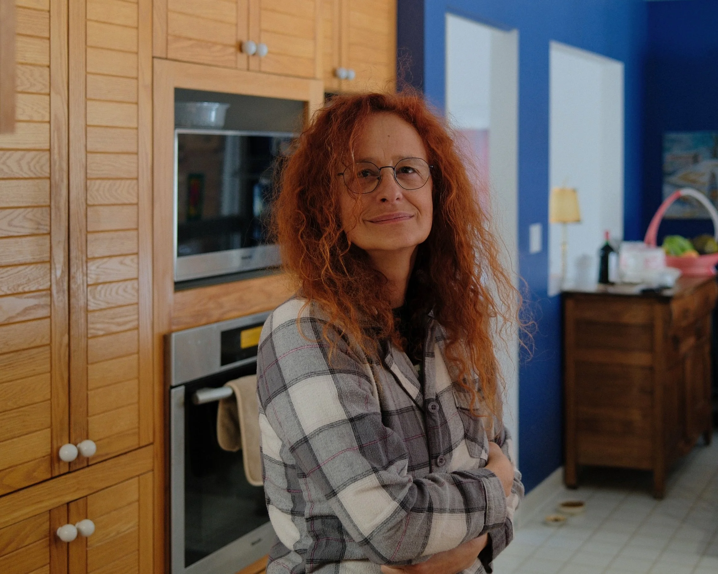 A woman with curly red hair, glasses, and a plaid shirt standing in a kitchen with wooden cabinets, a stove, and colorful walls.