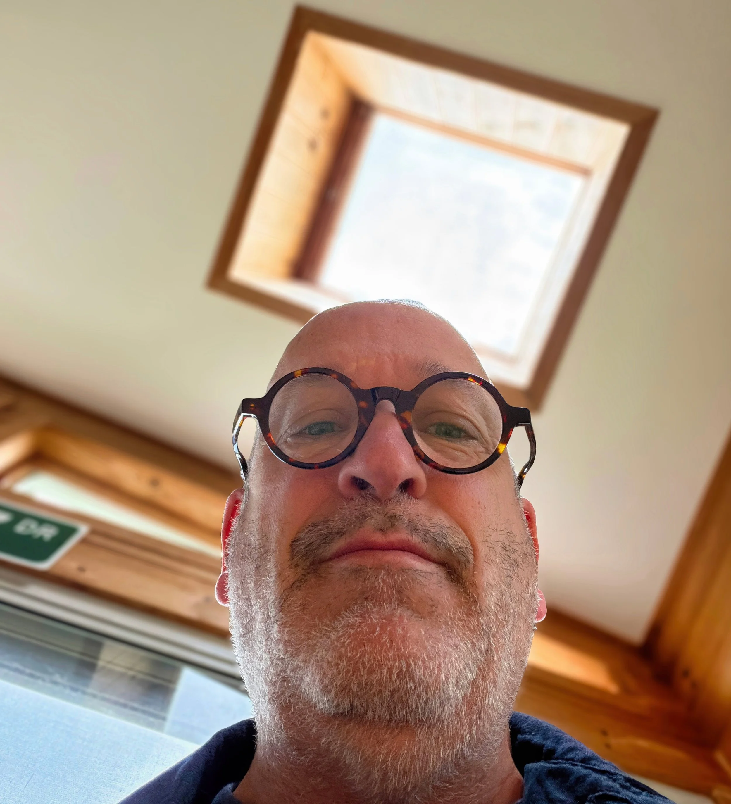 A person wearing tortoiseshell glasses has a close-up view of the top of their face, with the lower part of their face visible, including out-of-focus eyes, nose, and beard. The photo is taken indoors, showing a ceiling with a skylight and wood paneling.