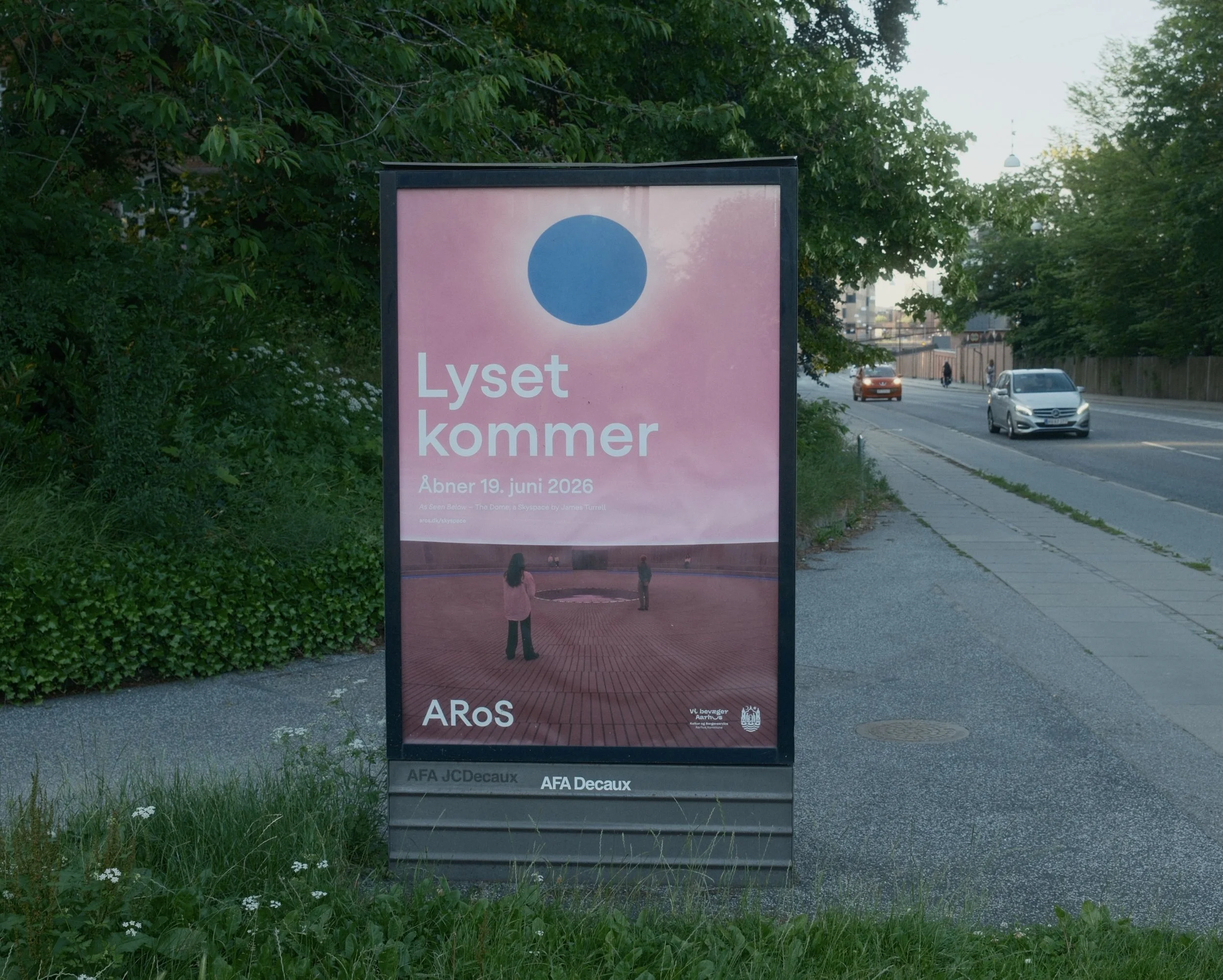 A digital billboard on a sidewalk displaying an advertisement for an art exhibition called 'Lyset kommer,' opening June 19, 2026, at ARoS museum, with greenery and cars on the street in the background.