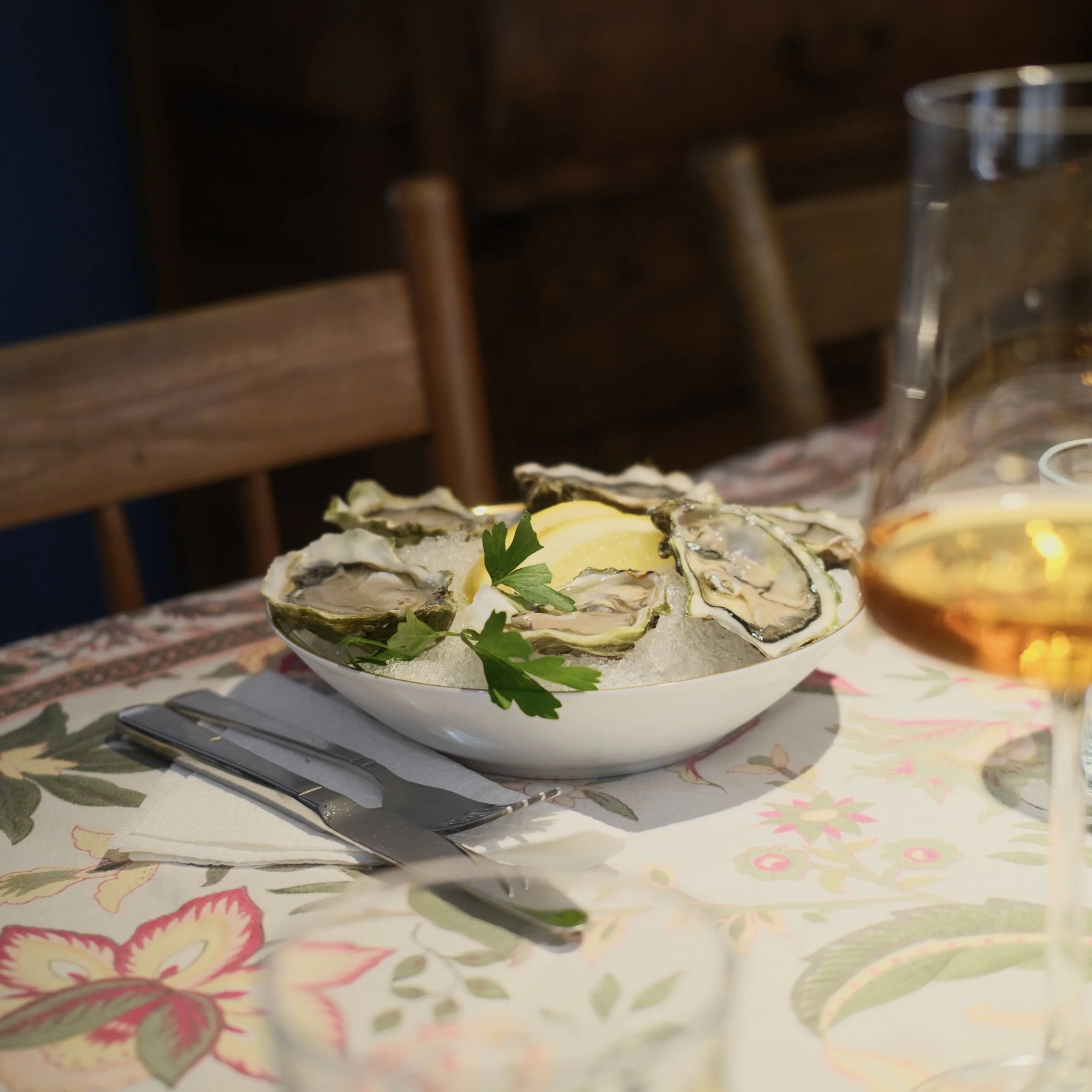 A plate of oysters garnished with parsley on an ornate tablecloth with a glass of water and some food items in the background.