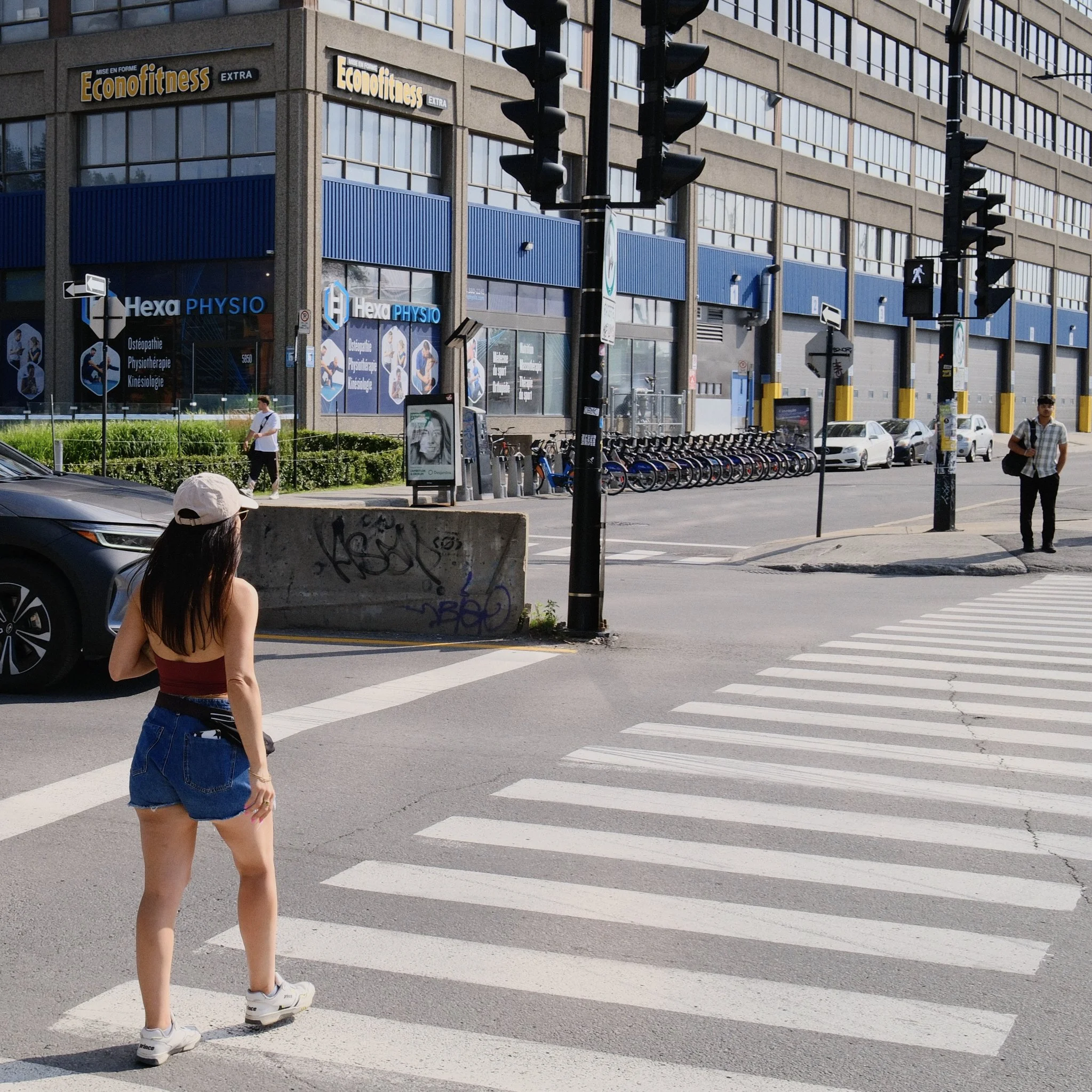 A young woman with long hair walking across a city crosswalk, wearing shorts and sneakers, with tall office buildings and traffic lights in the background.