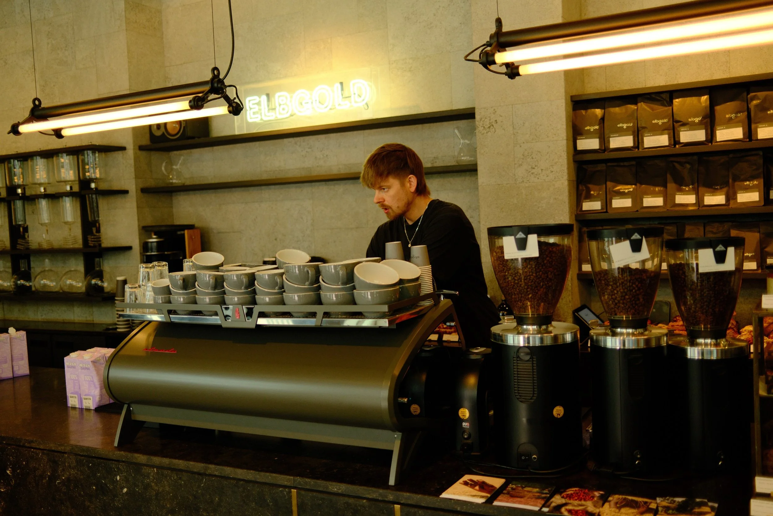 A barista working behind the counter of a coffee shop with rows of coffee beans and cups, and a neon sign that says 'ELBGOLD' on the wall.