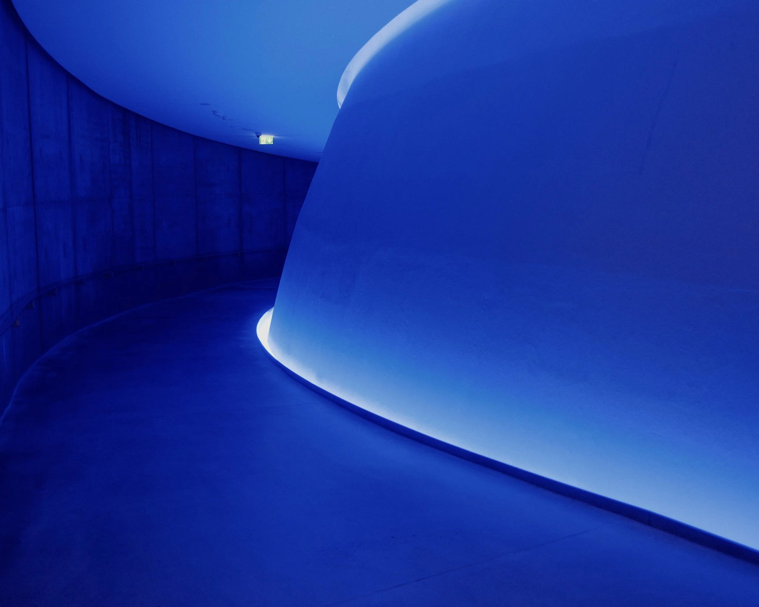 Blue abstract interior with curved walls and ceiling, lit by soft lighting, with an emergency exit sign in the distance.