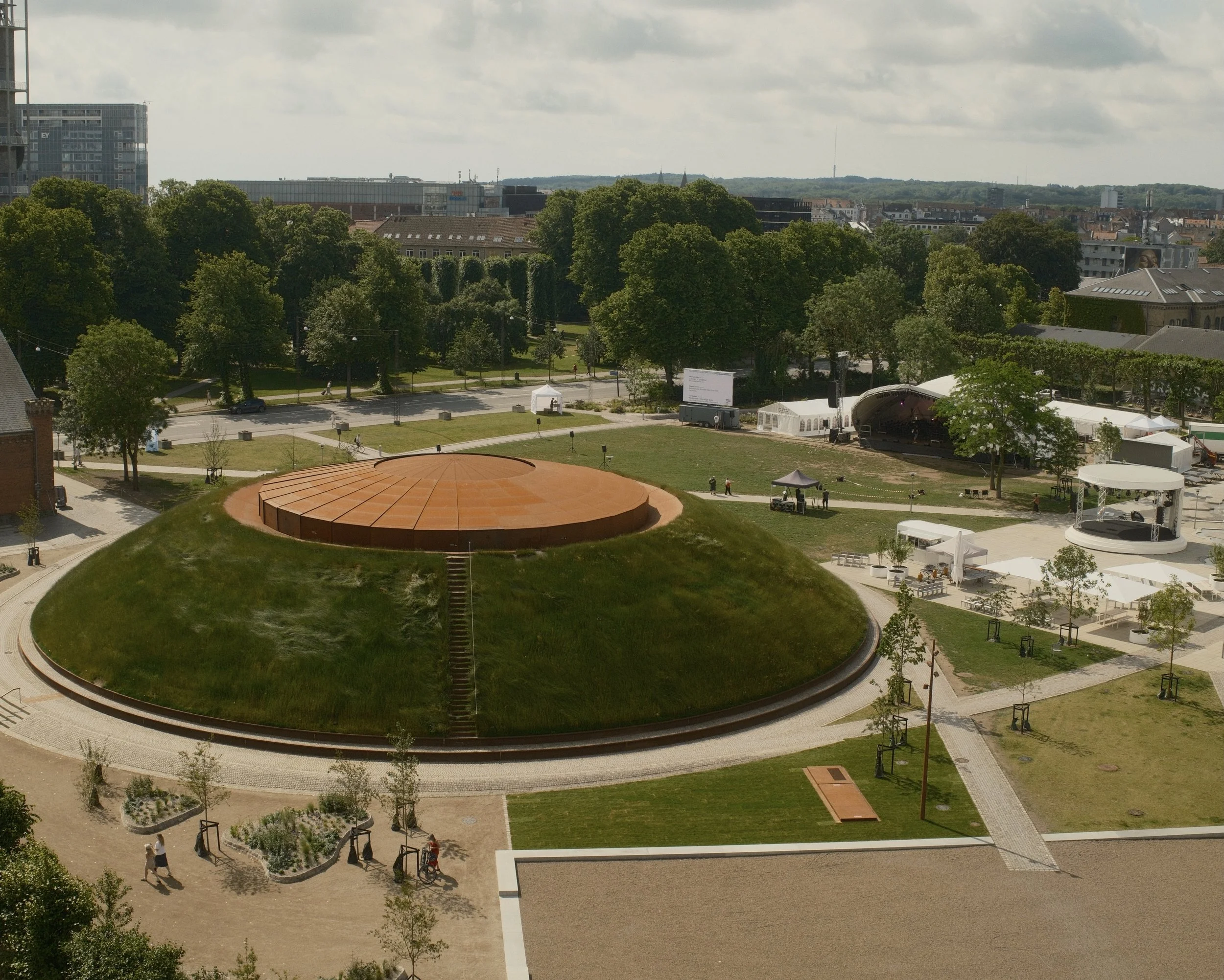A view of a park with trees and walking paths, outdoor tents, a stage, and people walking on the paths. In the background, there are buildings and a cloudy sky.