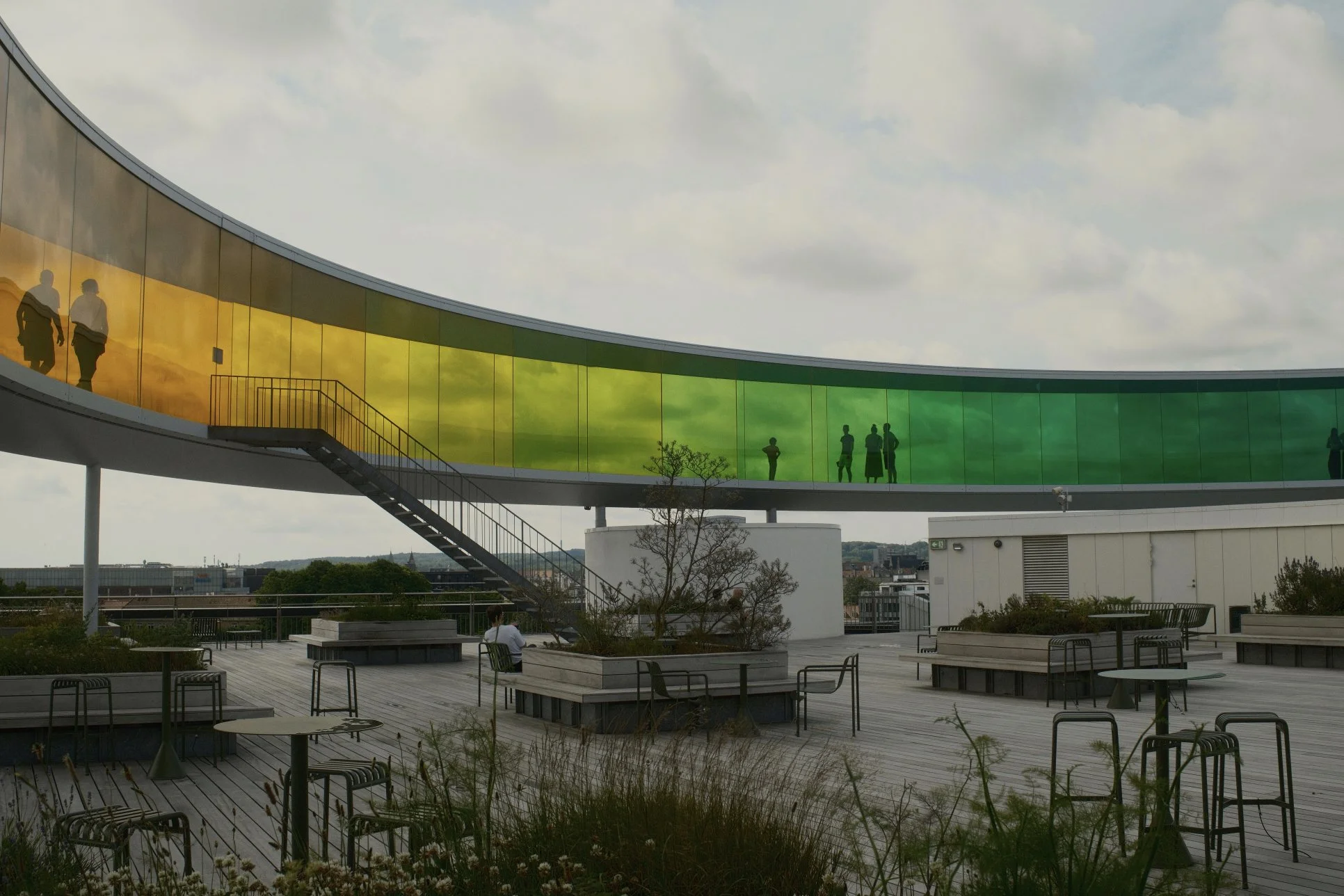 A rooftop observation deck with a curved, colorful glass walkway supported by white columns. Several people are walking along the walkway under a cloudy sky, and there are benches and tables with chairs on the deck.
