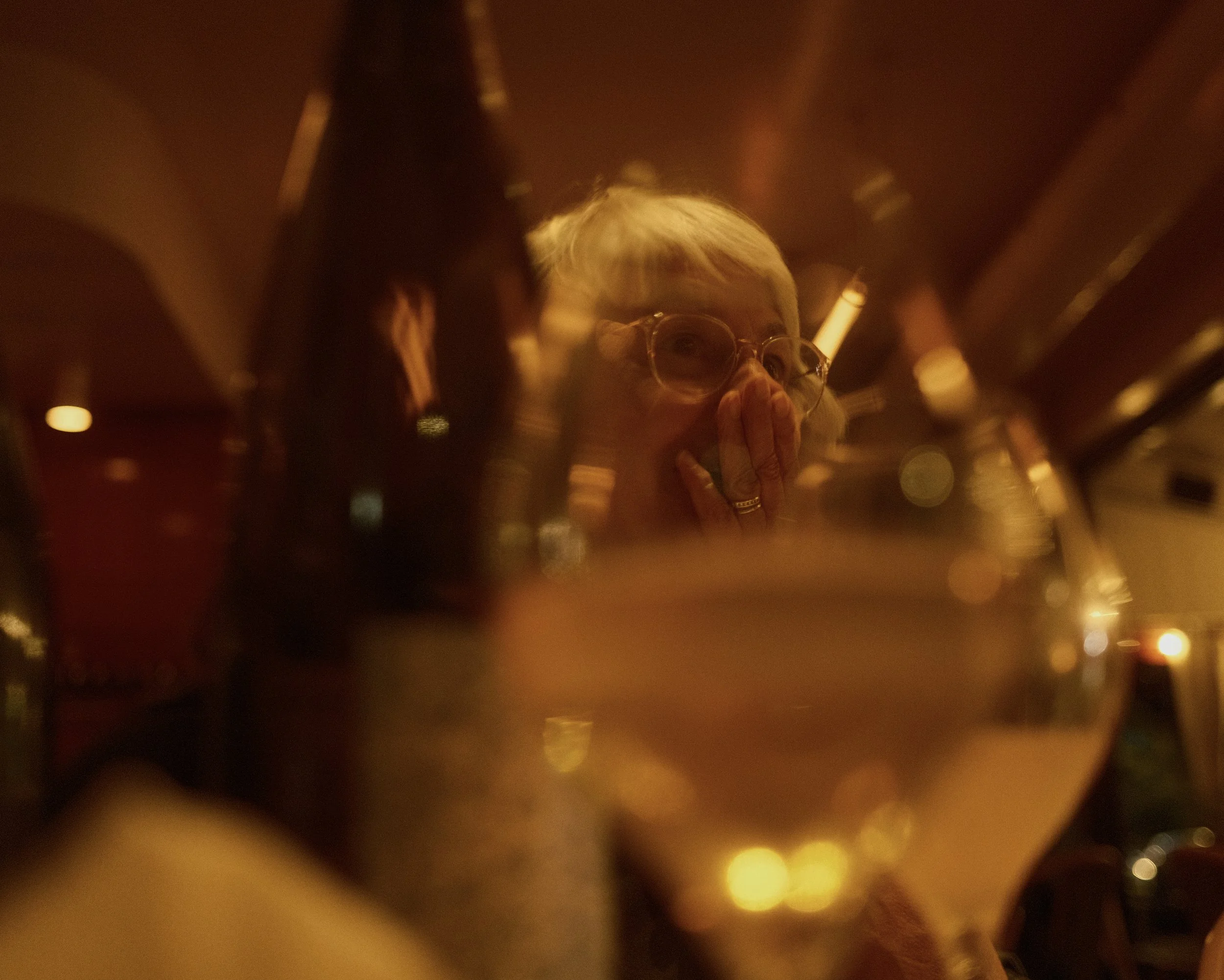 A woman with light hair and round glasses is seen covering her mouth with her hand in a dimly lit setting, possibly a restaurant or bar with warm lighting and out-of-focus background.