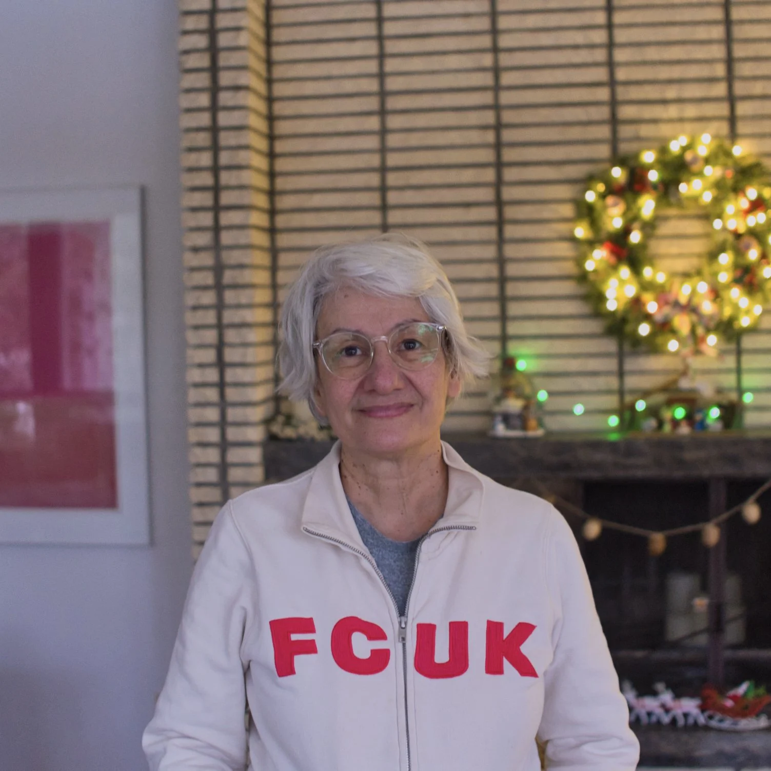 An elderly woman with short gray hair and glasses is smiling and standing in front of a fireplace decorated with a Christmas wreath and lights. She is wearing a white jacket with red letters that spell FCUK.