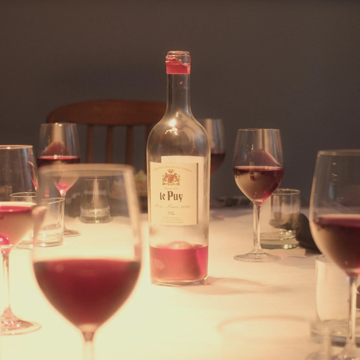 A bottle of wine labeled 'Le Puy' surrounded by several wine glasses filled with red wine on a table with a white tablecloth.