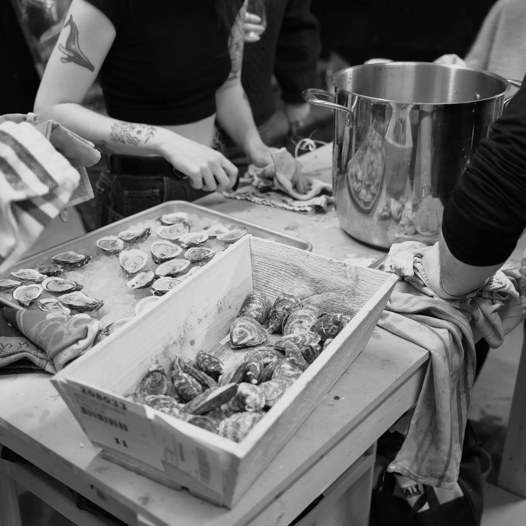 A person slicing smoked oysters on a cutting board. A box of oysters and a large metal container are on the table, with other people in the background.