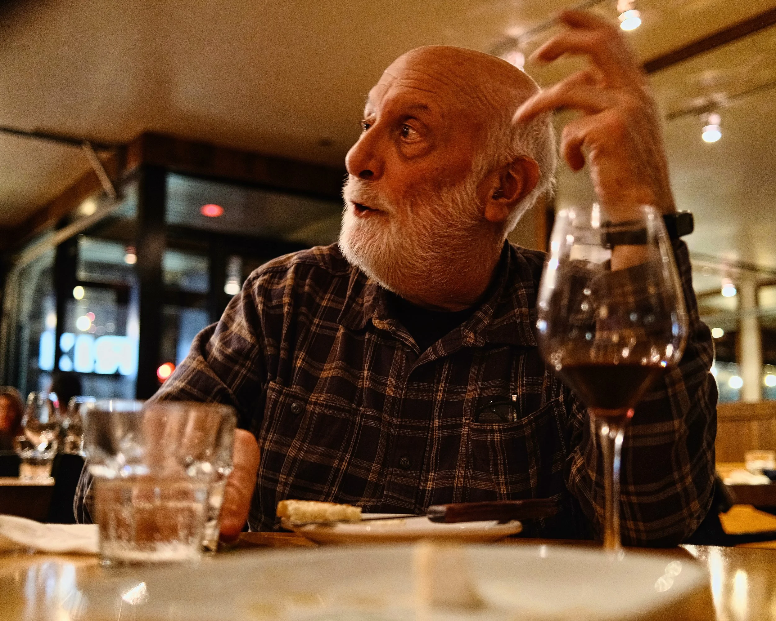 An elderly man with a white beard and plaid shirt sitting at a restaurant table, drinking from a glass of beer. The background shows a dimly lit dining area with warm lights.