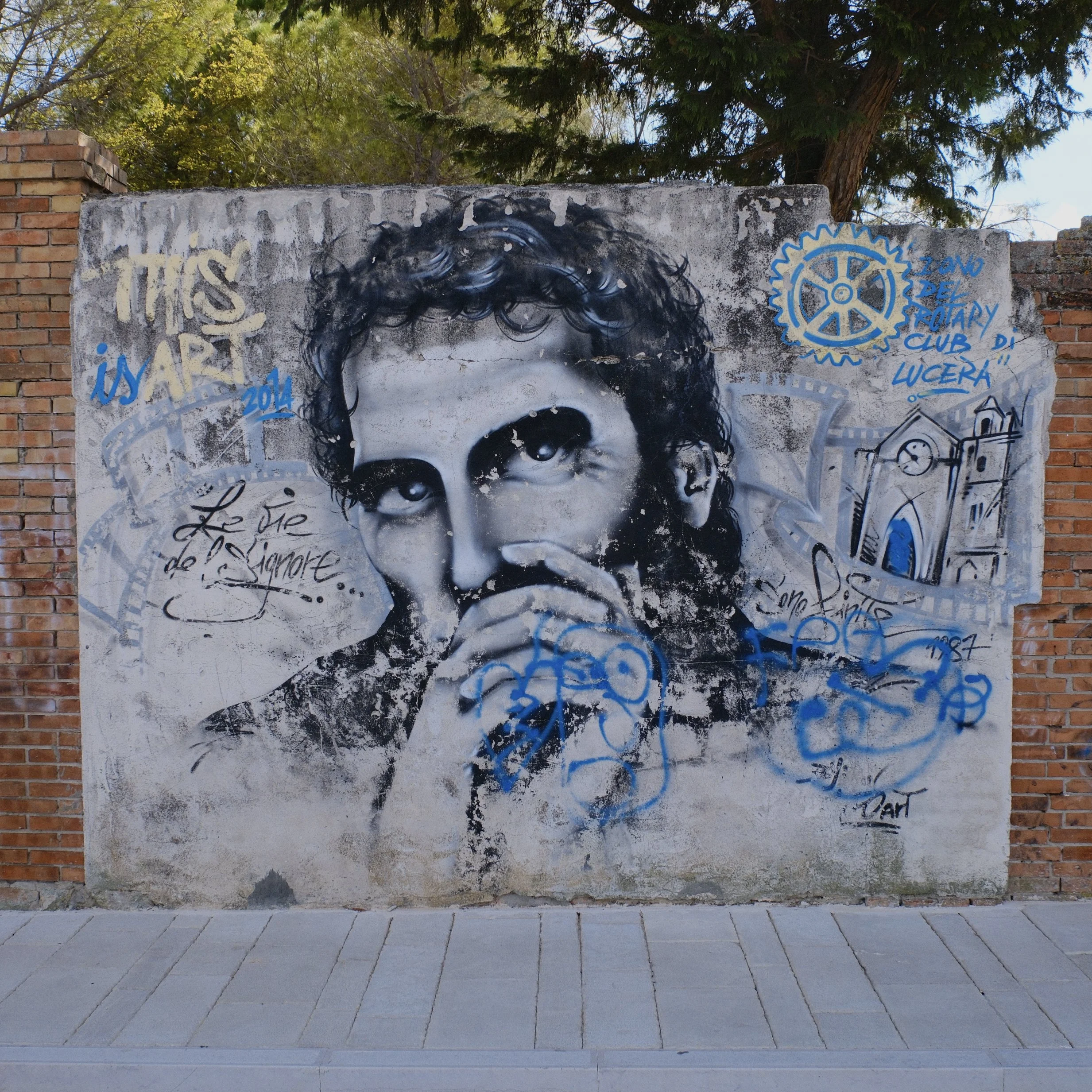 Graffiti mural on a concrete wall depicting a stylized black and white portrait of a young girl with curly hair, alongside blue and yellow text and symbols, including the Rotary Club logo, connects to a brick wall and green trees in the background.