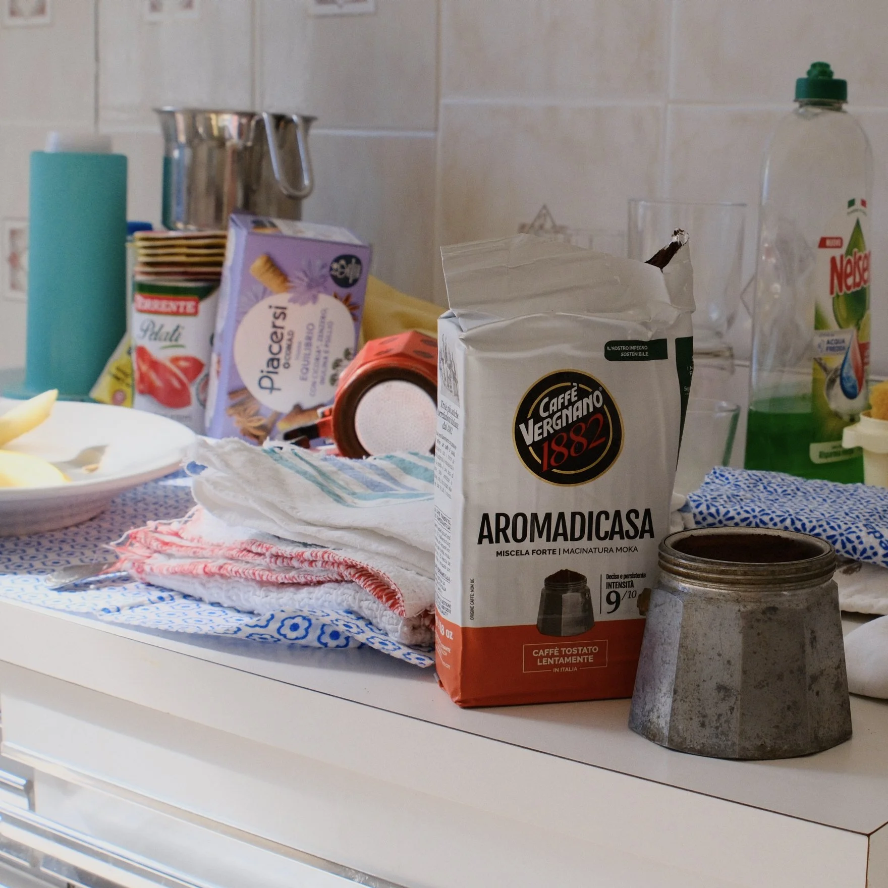 A collection of everyday items on a kitchen counter, including a coffee bag labeled "Aromadicase," a box of tea, a glass of water, a water bottle, a container of tomato sauce, and some assorted utensils and cloths.