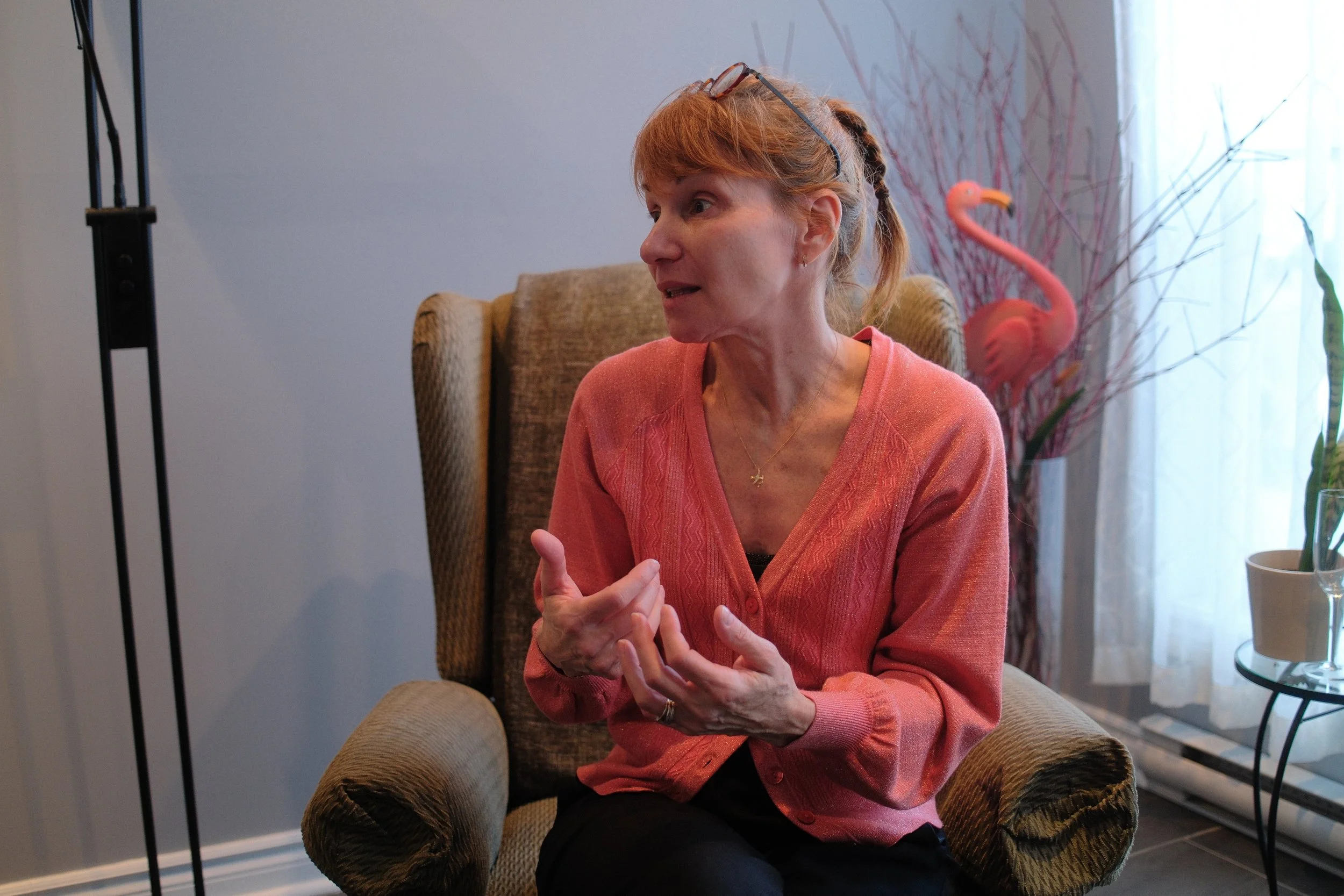 A woman with curly red hair, glasses on her head, wearing a pink sweater, sitting in an armchair, gesturing with her hands, in a room with decorative plants and a window with curtains.