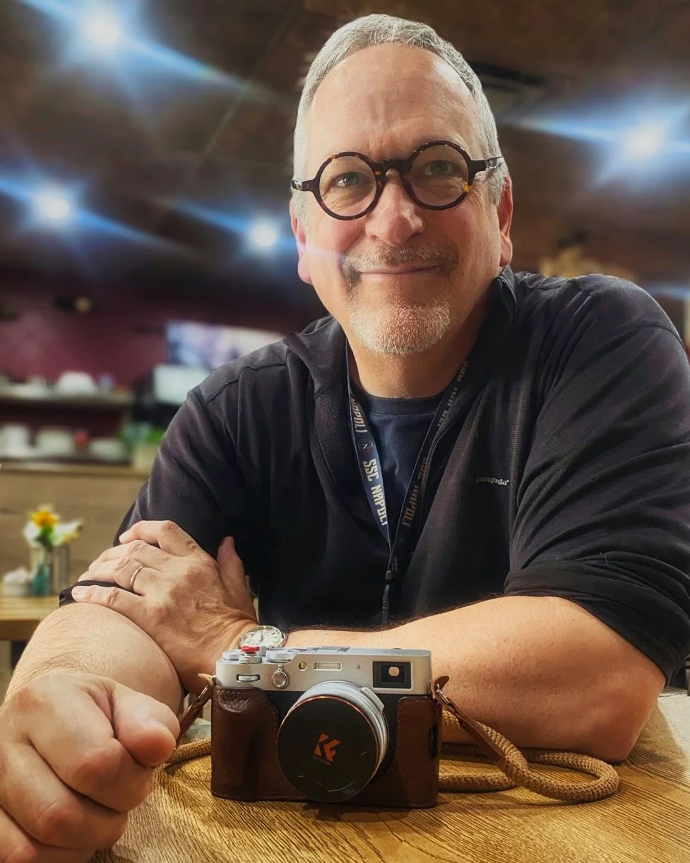 A smiling middle-aged man with glasses and a beard sitting at a wooden table with a vintage camera in front of him. He is wearing a black jacket and a lanyard around his neck. The background shows a warm, dimly-lit indoor setting.