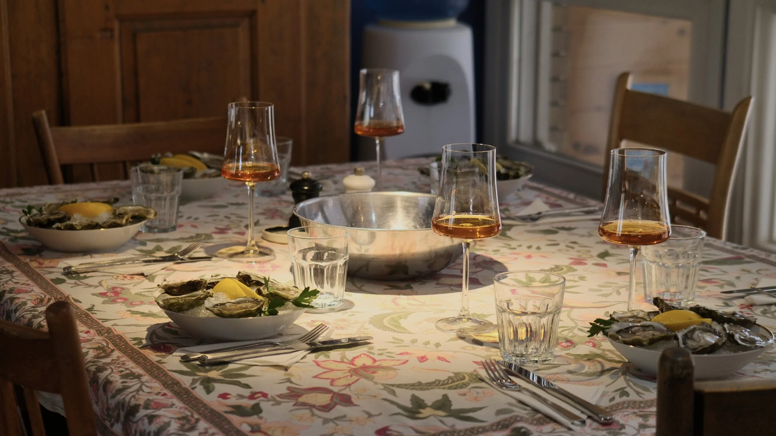 A dinner table with open oyster shells, lemon wedges, and parsley, set with forks and glasses of rosé wine, in a cozy wooden dining room.