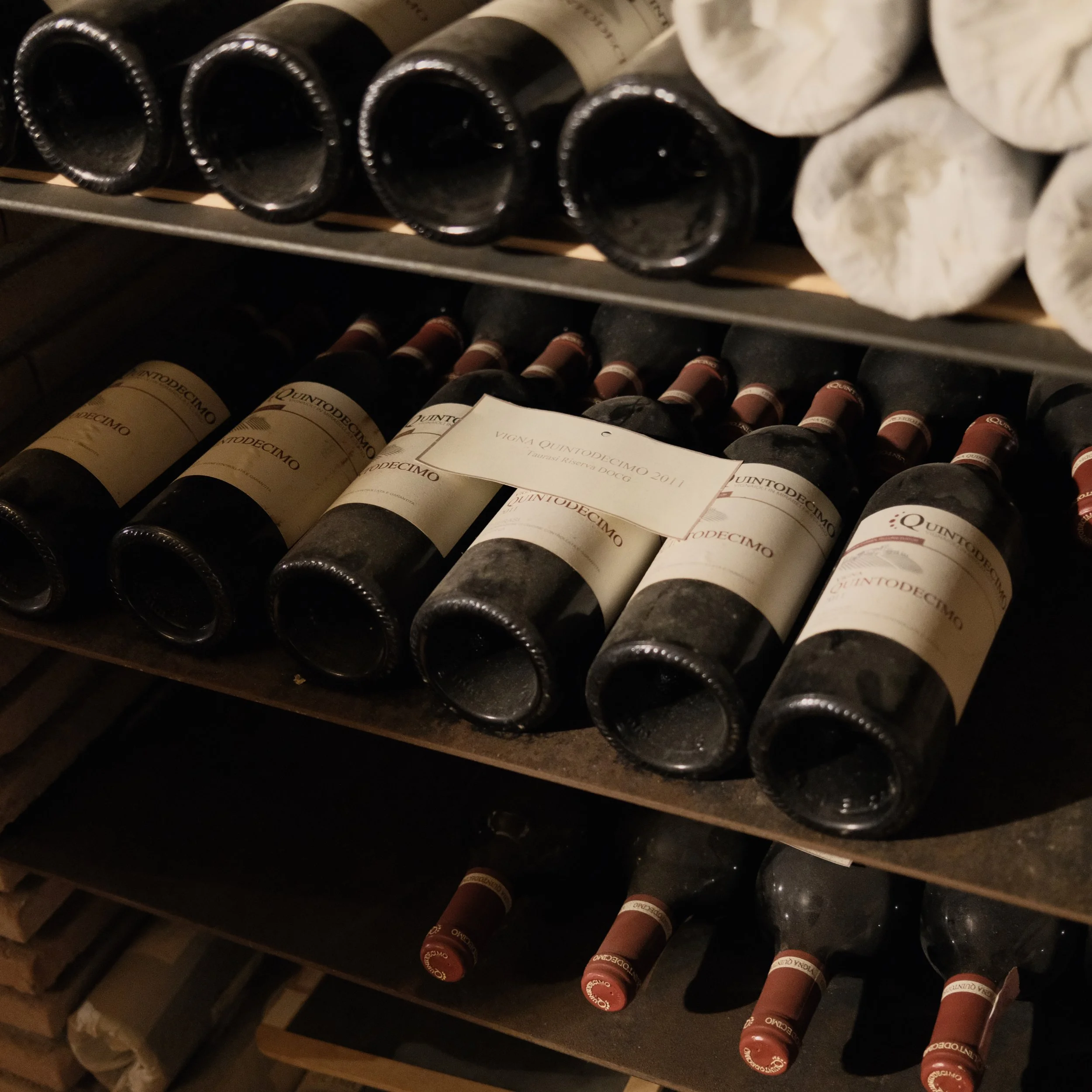 A wine rack holding several bottles of wine, with red wine bottles in the bottom row and other bottles on the upper row labeled Quintodecimo.