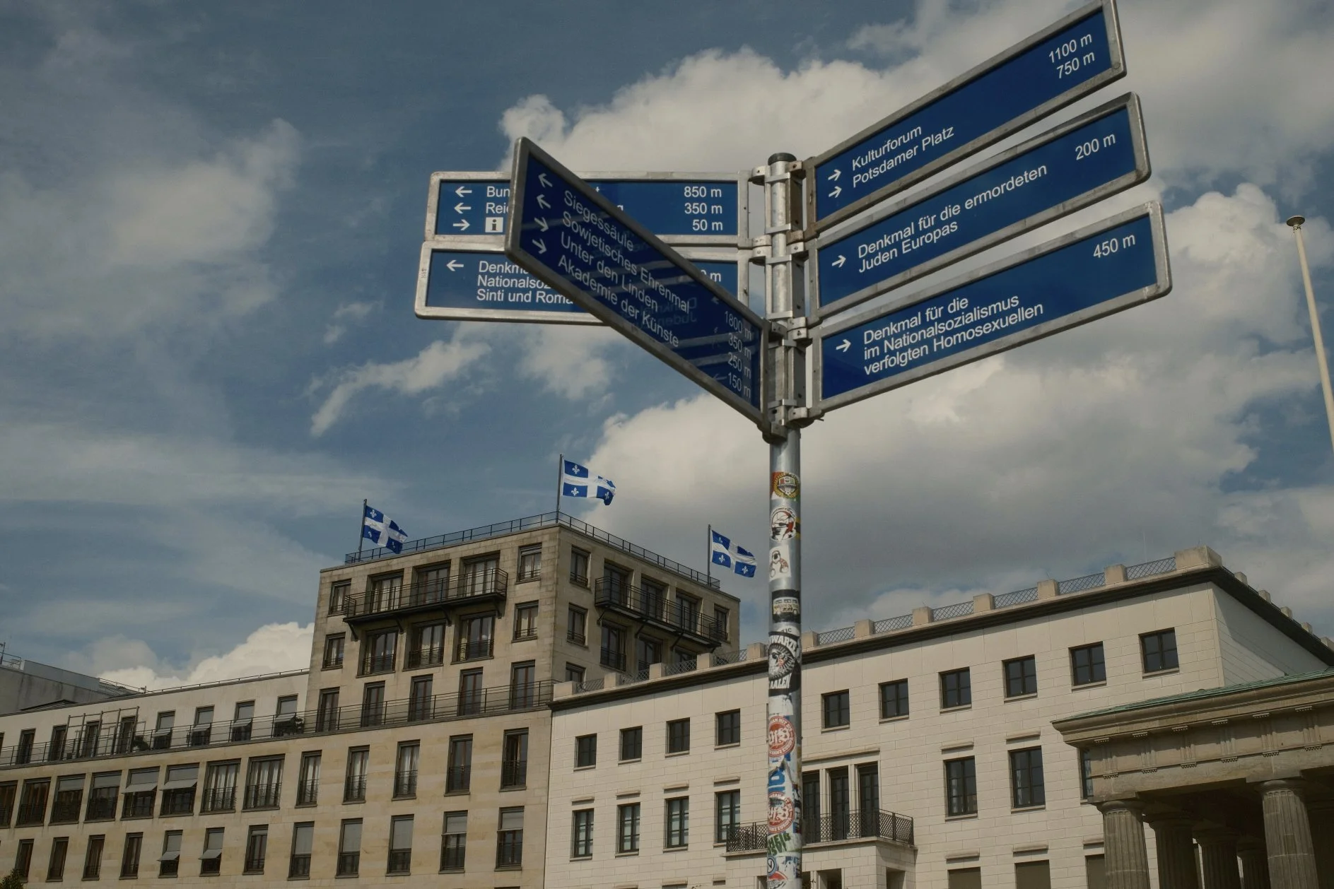 Street sign with multiple blue directional panels in German, mounted on a pole covered with stickers, in an urban area with a beige multi-story building and flags in the background, under a partly cloudy sky.