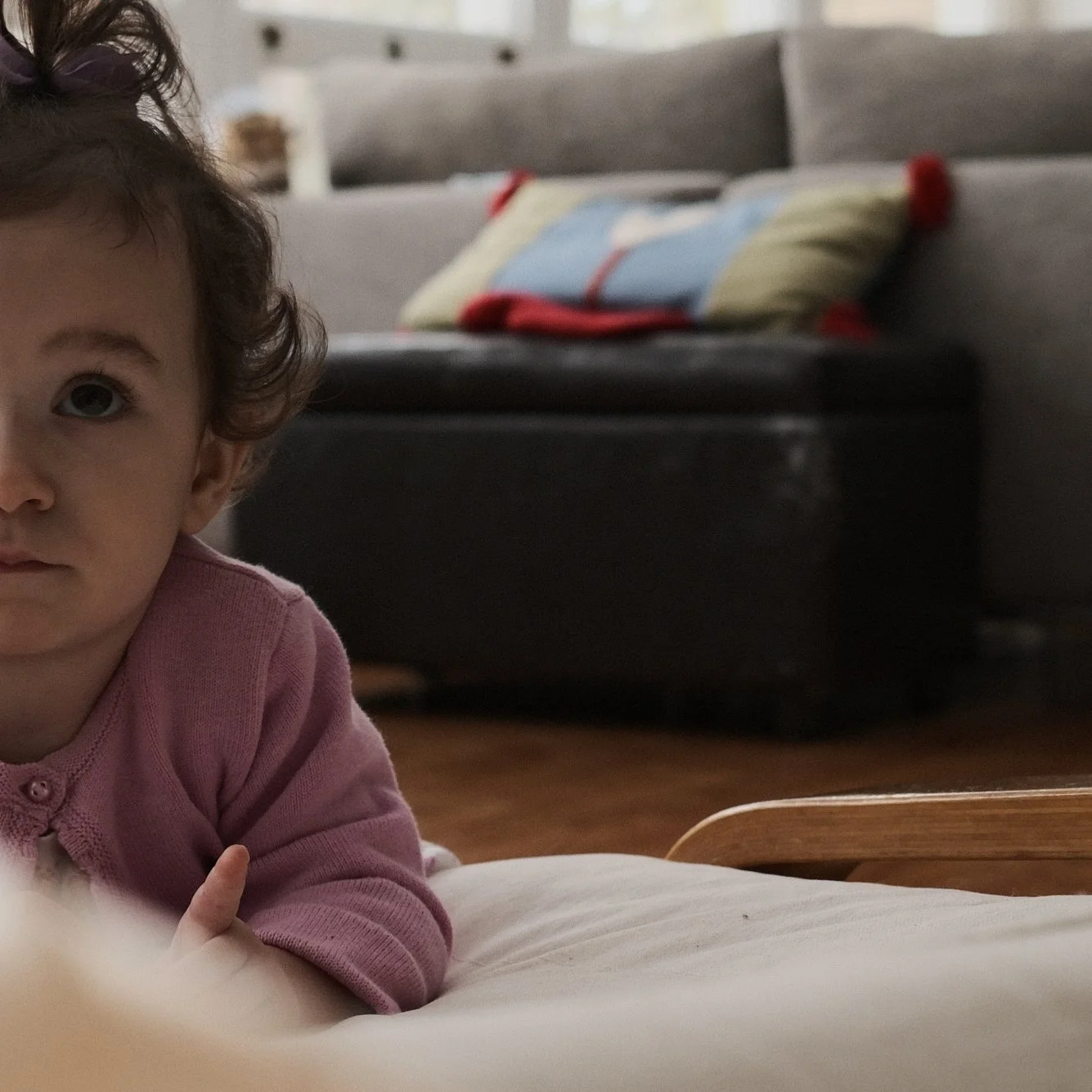 Close-up of a young girl with curly brown hair, wearing a pink top, lying on a bed with a wooden headboard, partially visible in a bedroom with a gray couch and a colorful pillow in the background.