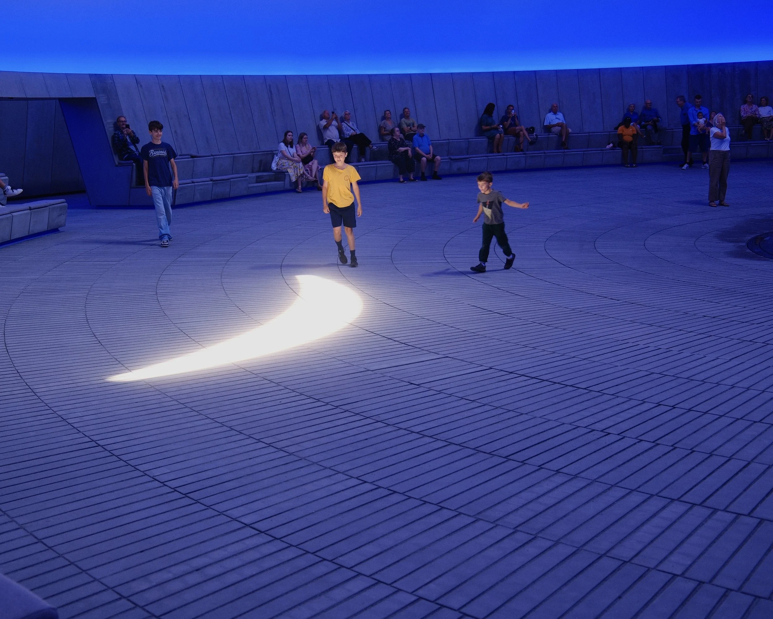 People sitting on blue benches in an indoor space with blue lighting, some people standing, children walking across the floor.