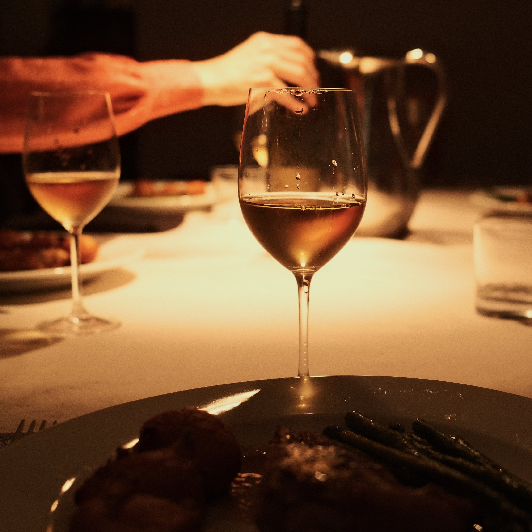 Close-up of a wine glass filled with white wine, with another glass and a partially visible plate in the background. The scene is dimly lit, creating a warm, cozy atmosphere.