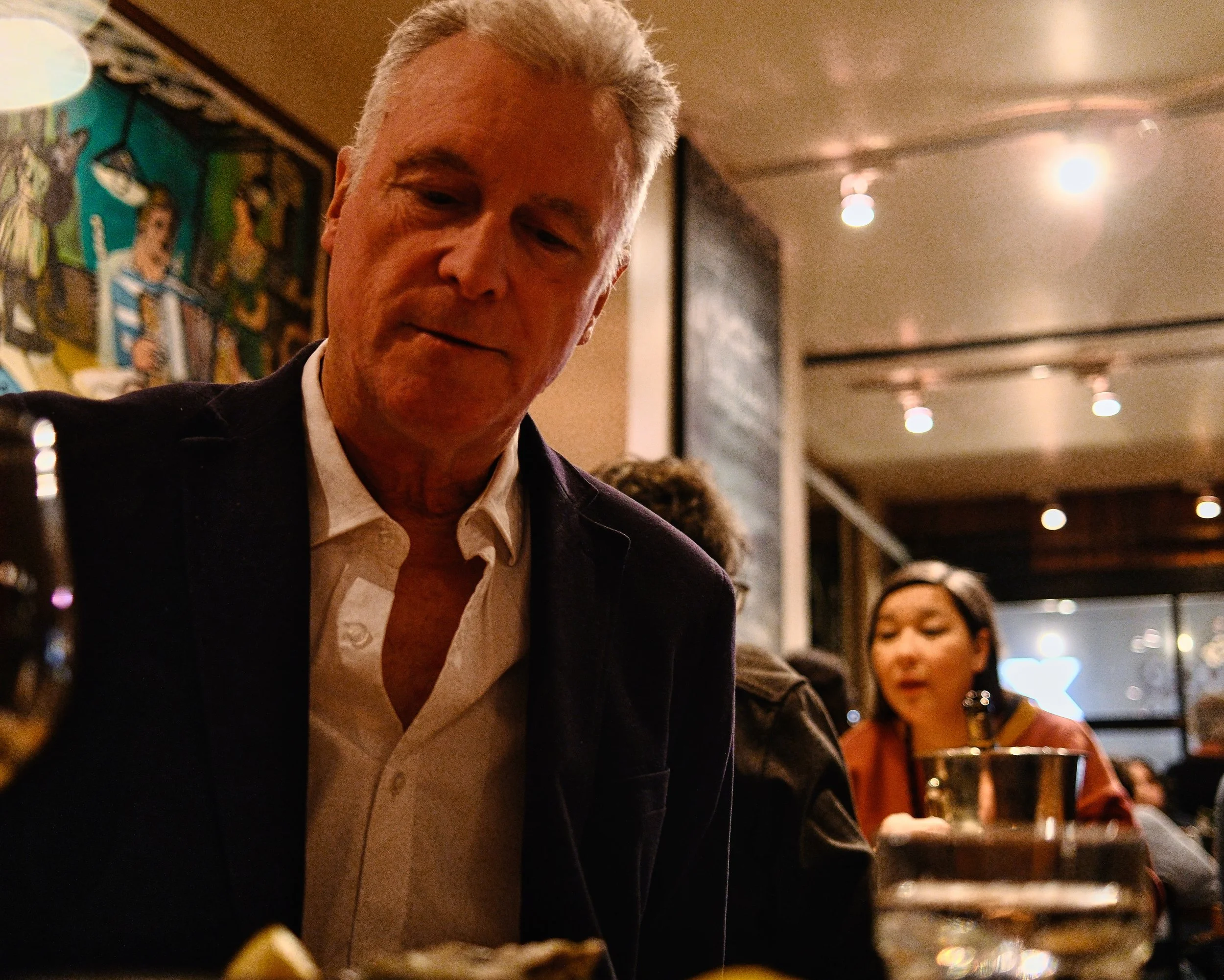 A man with gray hair dressed in a white shirt and dark blazer is seated at a table in a dimly lit restaurant or bar. A woman with dark hair and reddish shirt is visible in the background, sitting at the same table with a drink in front of her. The de
