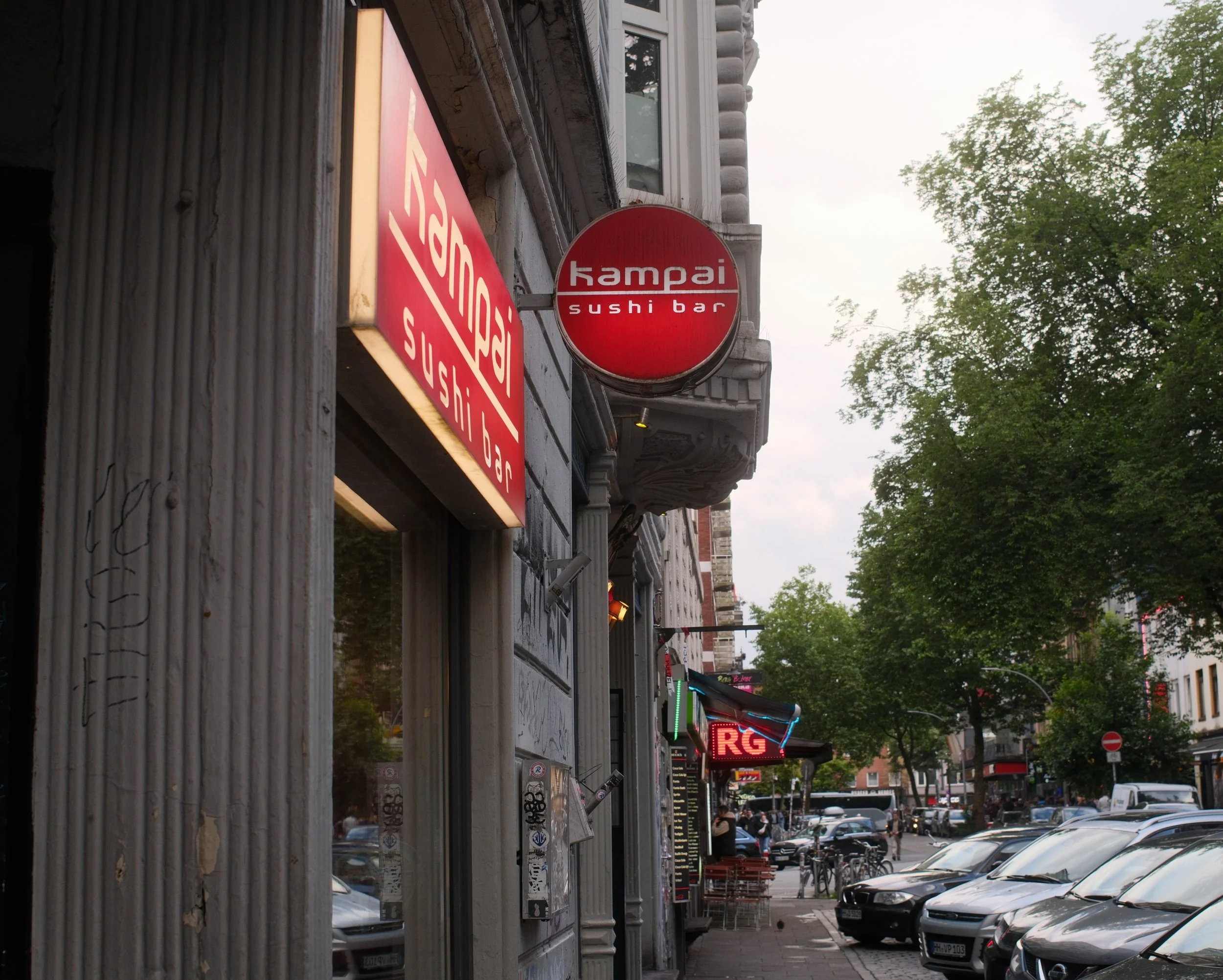 Street view featuring a sushi bar named 'Kampai' with a red circular sign, other storefront signs, parked cars, pedestrians, and trees lining the street.