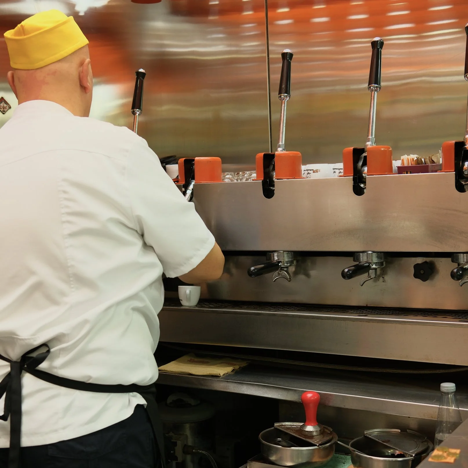 A chef in a white uniform and yellow hat seen from the back behind a stainless steel counter with cooking utensils and equipment in a restaurant kitchen.