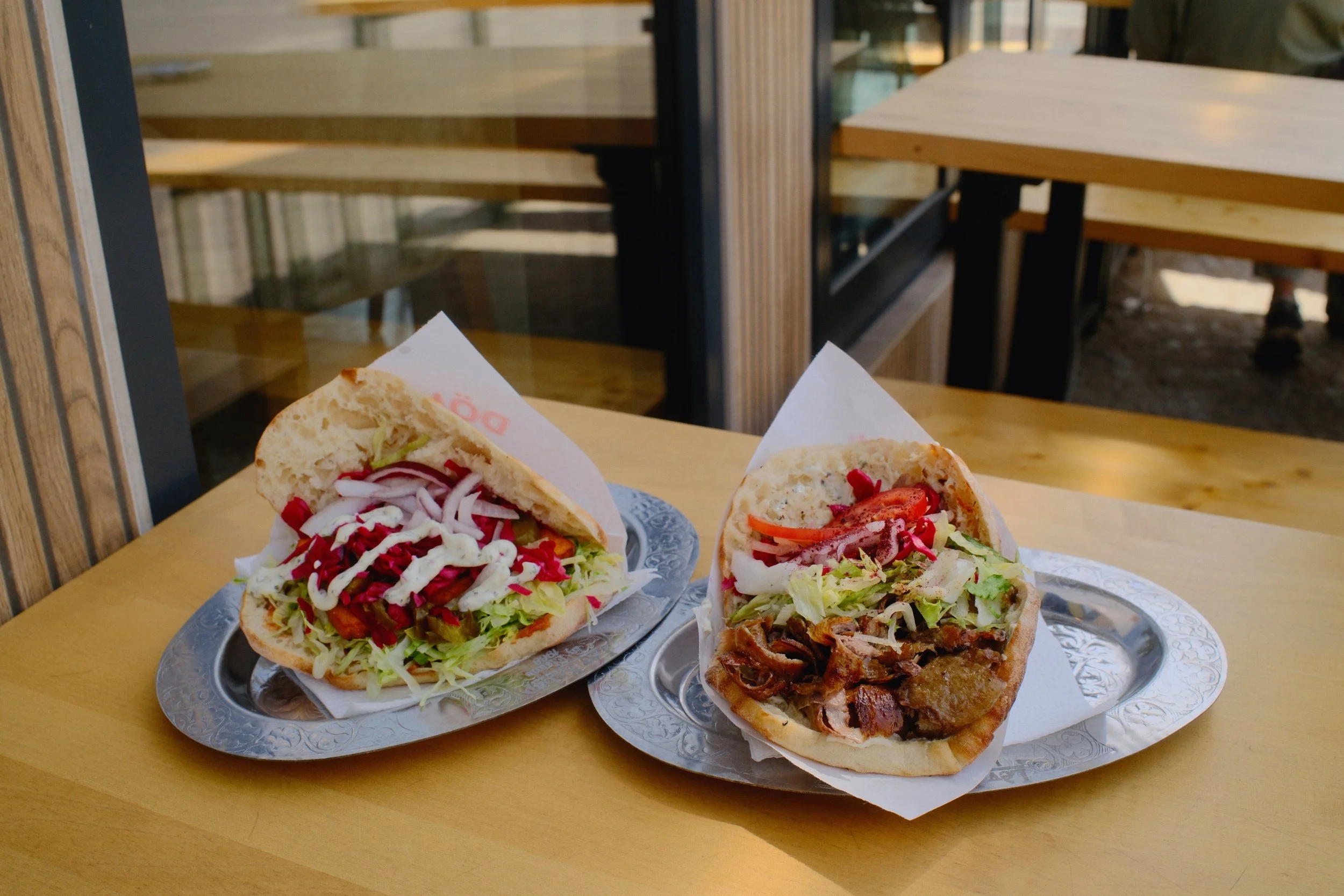 Three pita sandwiches filled with vegetables and meat, served on metallic trays on a yellow table inside a restaurant.