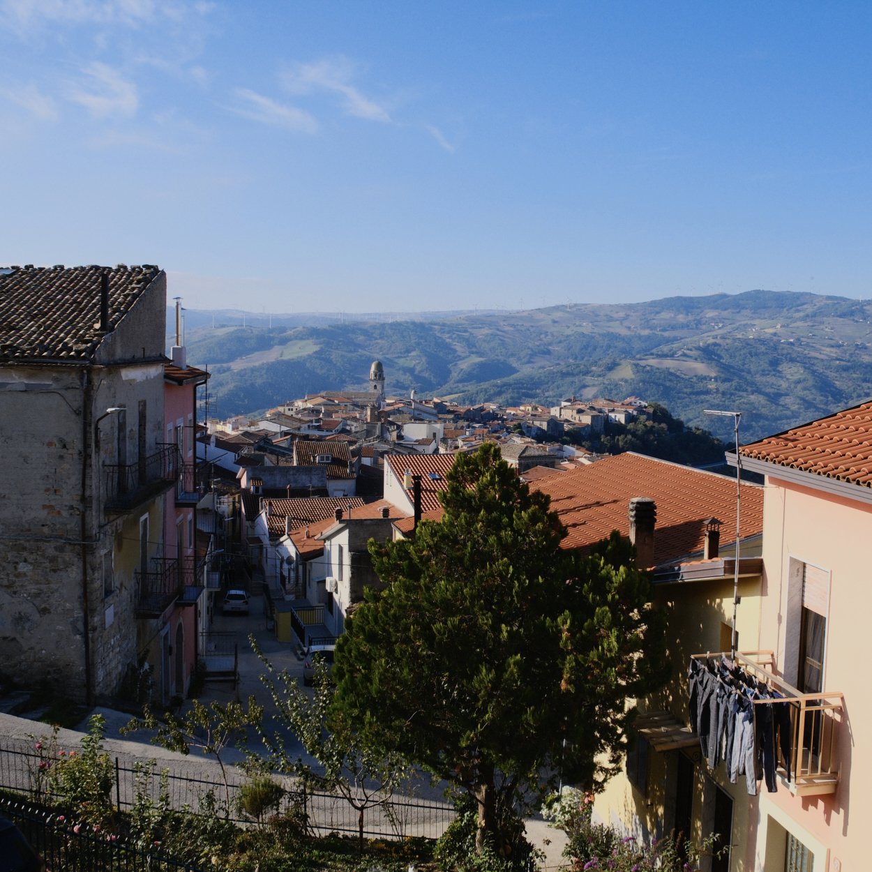 Hilly Italian town (San Bartolomeo in Galdo) with colorful buildings, red-tiled roofs, and mountains in the background under a blue sky.