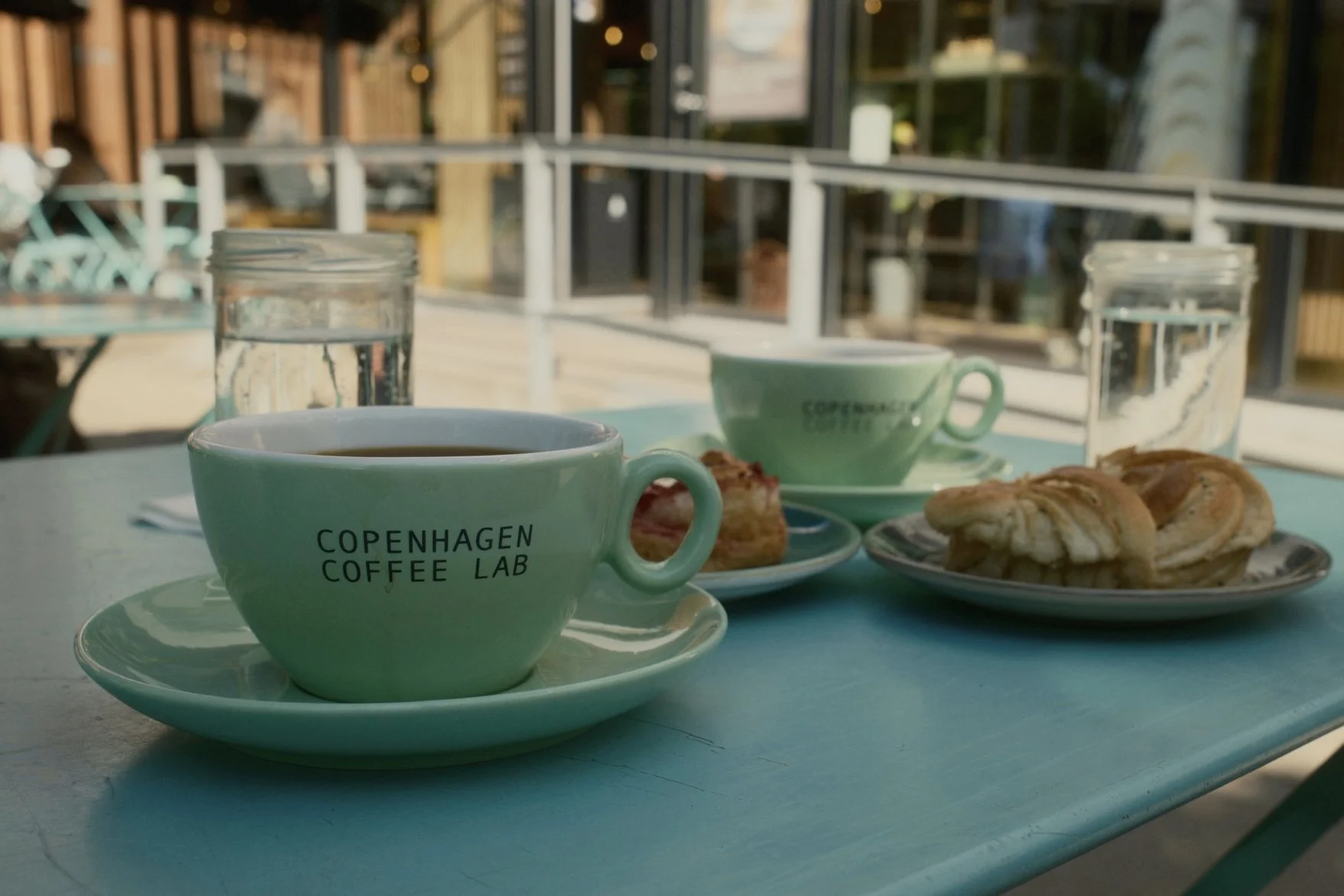 Two mint green coffee cups with the text 'Copenhagen Coffee Lab' sit on a white table, with plates of pastries and two glasses of water. In the background, a balcony and glass door are visible.