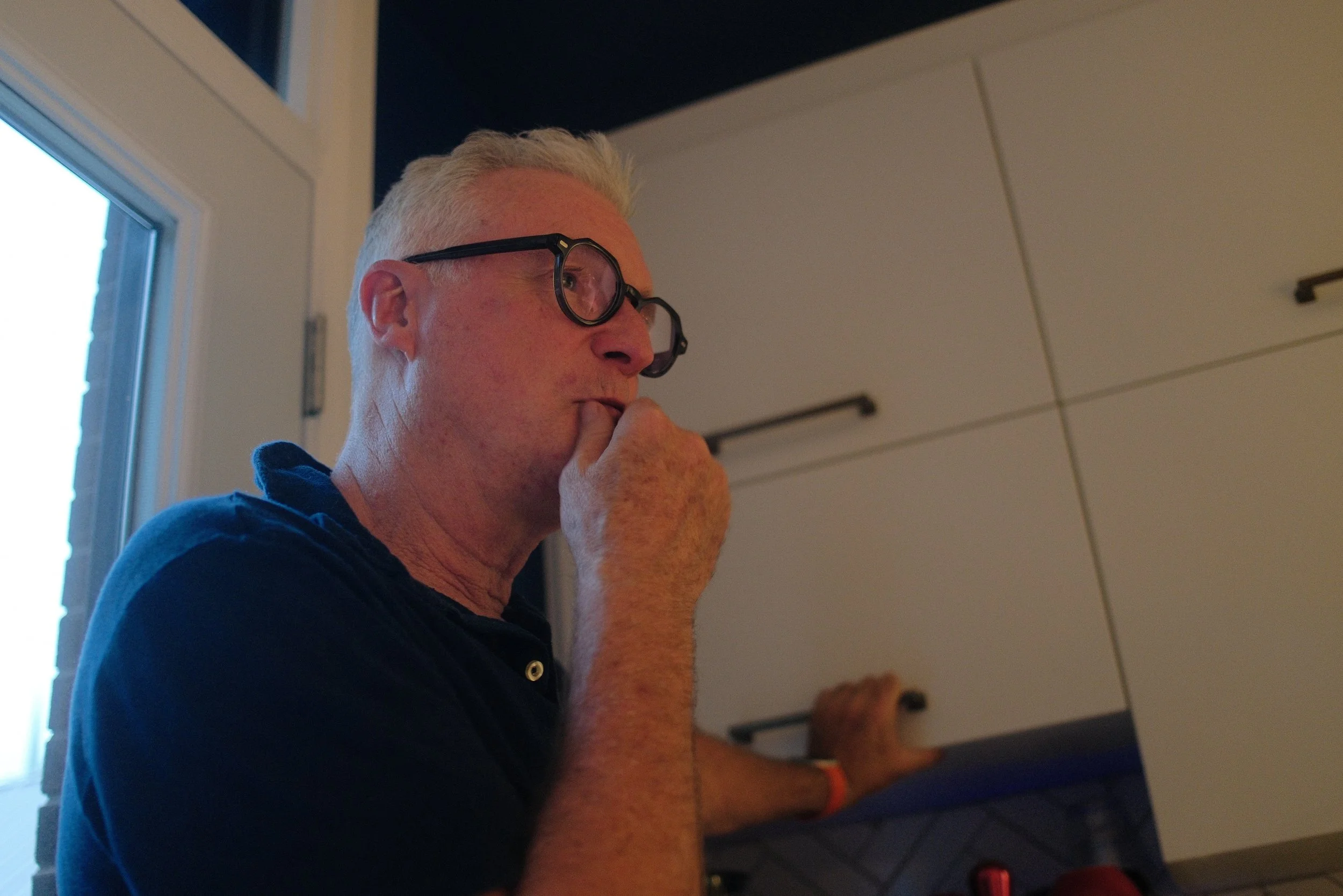 A man with short, white hair and glasses is sitting near a window, holding his chin with his hand, appearing to be in thought.