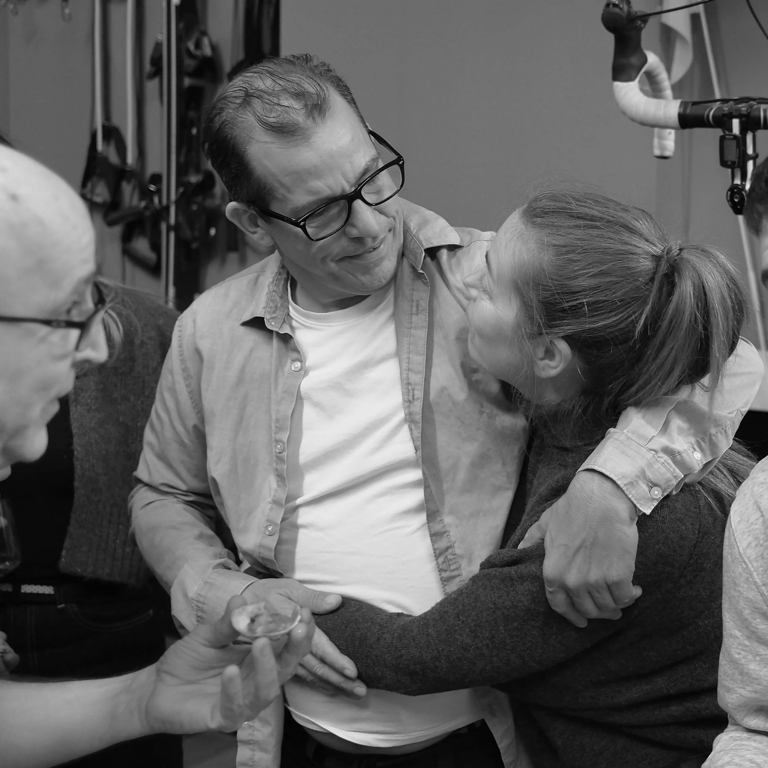 A man wearing glasses holds a small plastic cup in his left hand and appears to be looking down at something out of view, while someone else's hand gently touches his shoulder. The scene takes place in a clinical or laboratory setting with equipment visible in the background.