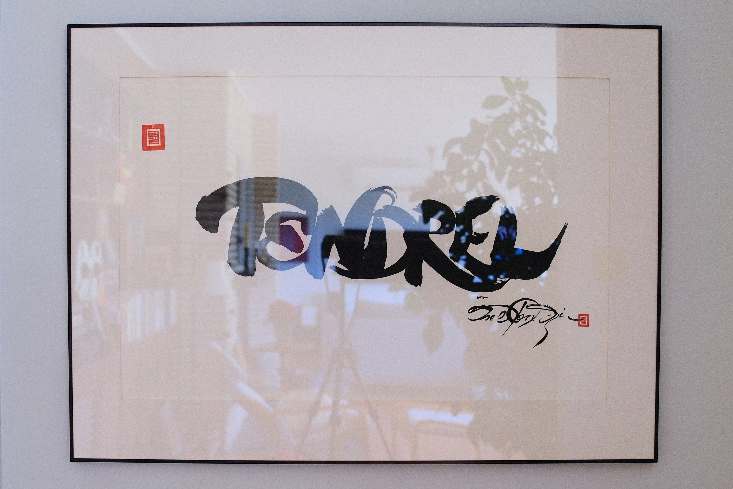 A framed piece of abstract Asian calligraphy art with black brush strokes on white paper, featuring red seals in the upper left and bottom right corners, hanging on a white wall.