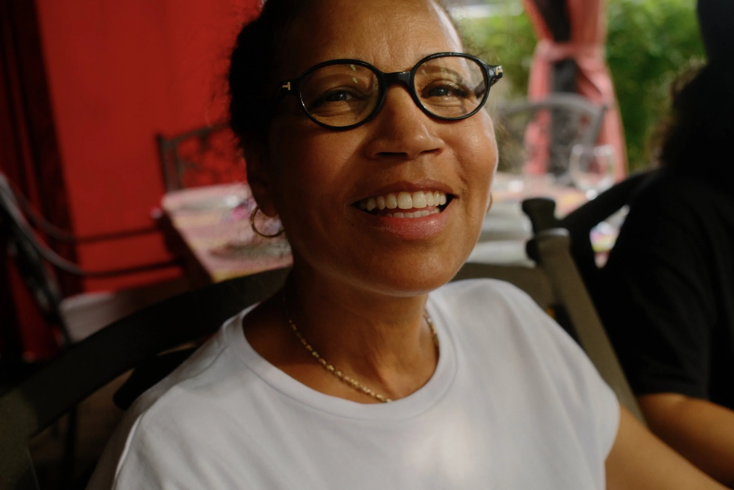 A woman with glasses smiling and wearing a white shirt, with a red background and outdoor greenery in the background.