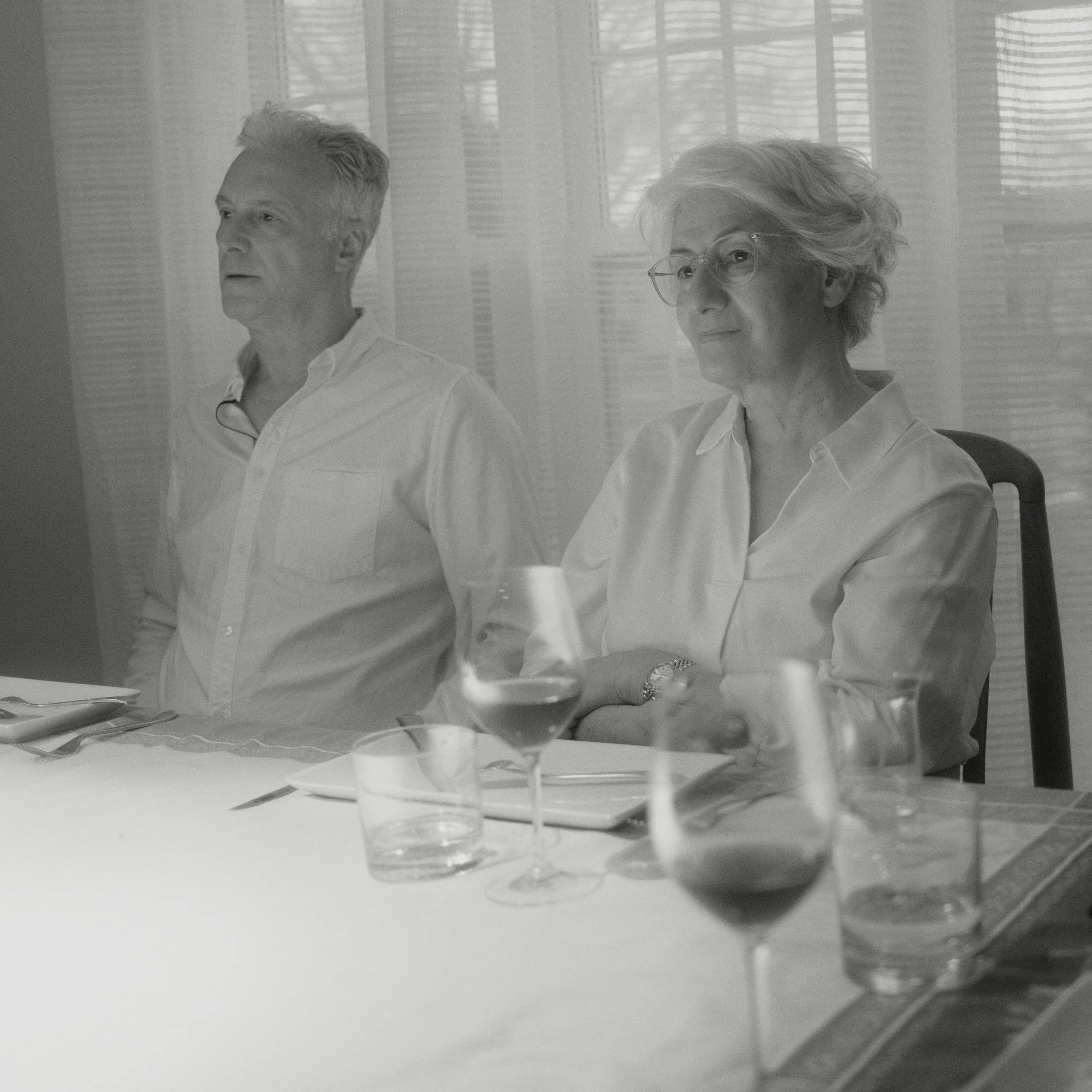 A grayscale photograph of two elderly women seated at a table with glassware and plates, in front of window blinds.