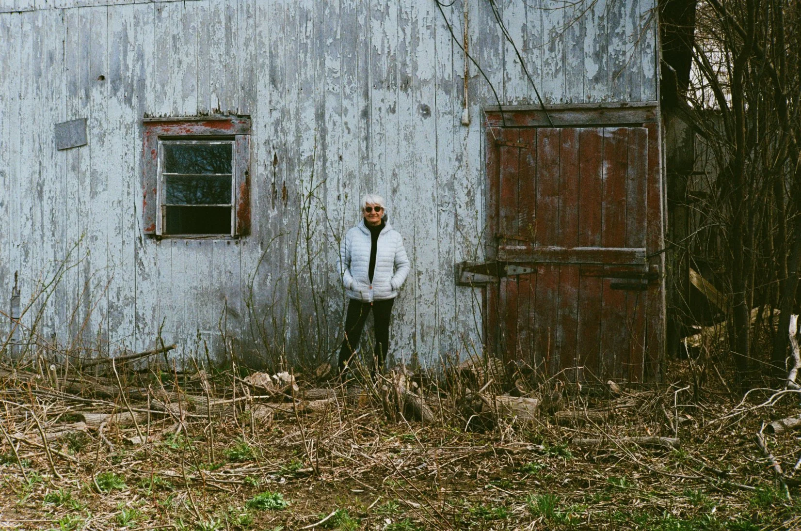 Woman standing in front of an old, weathered barn with peeling paint and a closed sliding door, surrounded by overgrown vegetation.