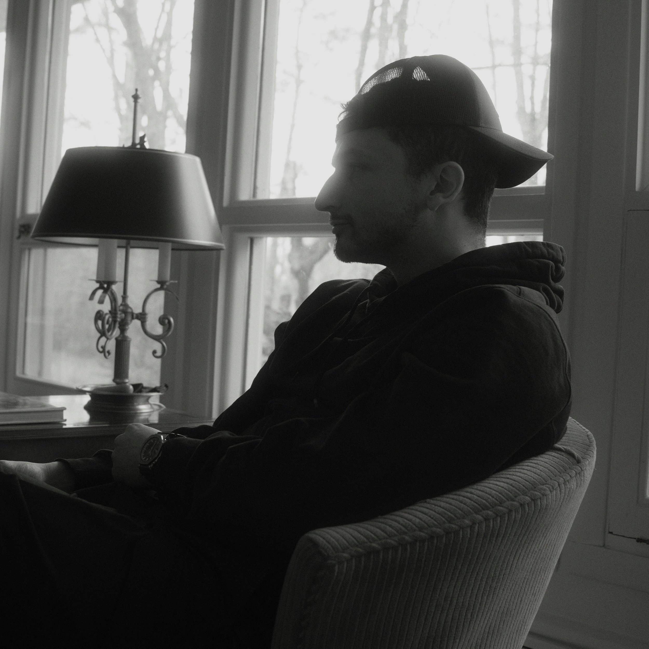 Side profile of a man sitting indoors, wearing a baseball cap, with a window and tree branches visible in the background.