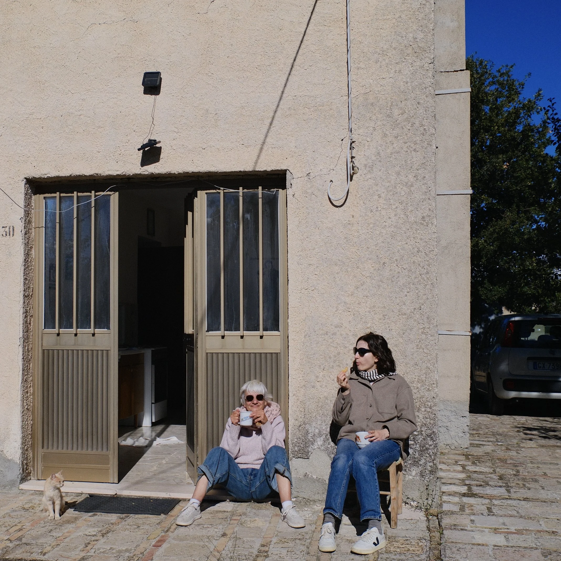One elderly woman and a younger woman sitting outside a house with a concrete wall, a window, and a door. They are enjoying drinks and a cat is sitting nearby. The older woman is sitting on the floor and the younger woman is sitting on a chair and ea