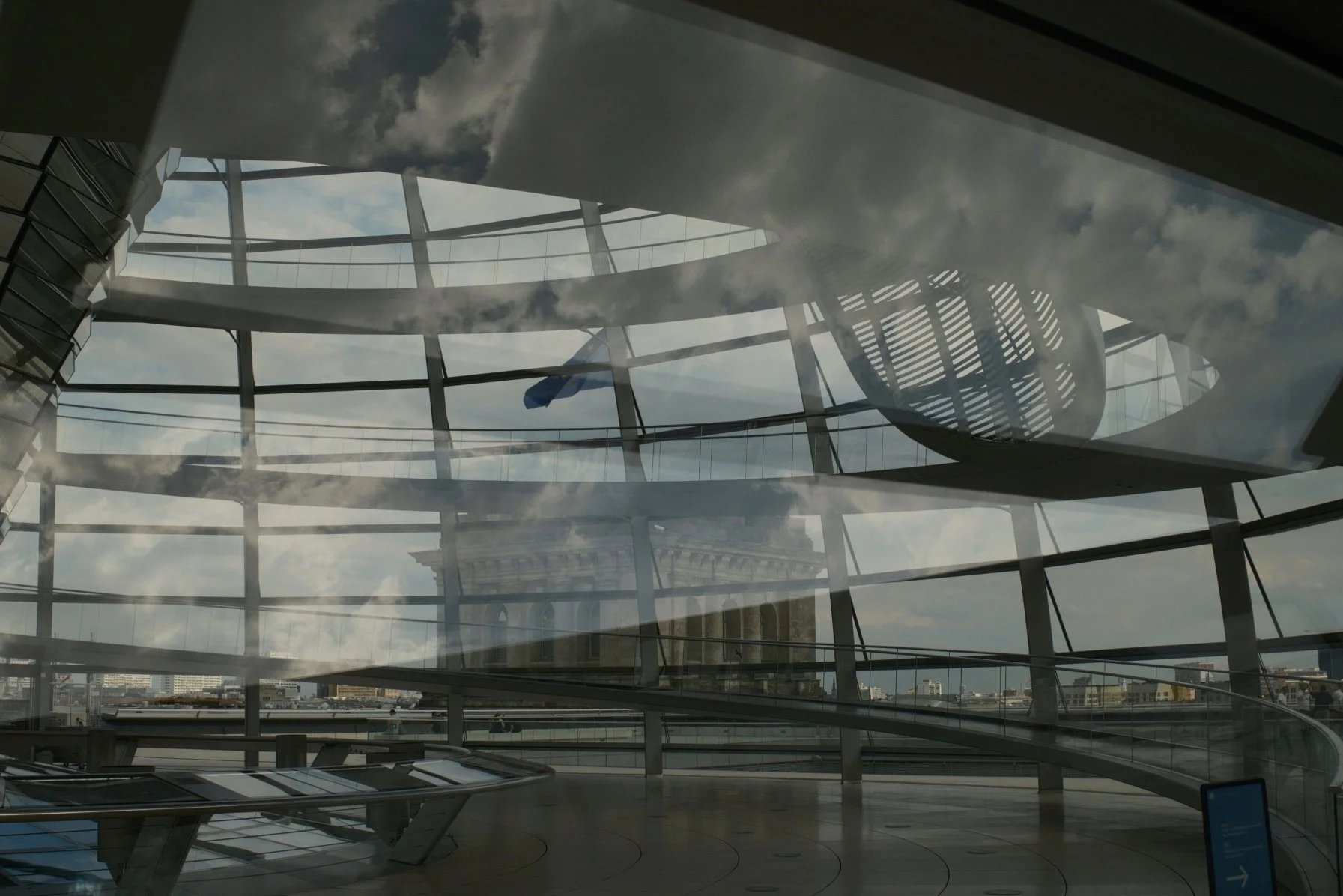 View of a modern glass ceiling interior with reflections of the sky and clouds, and a flag visible outside.
