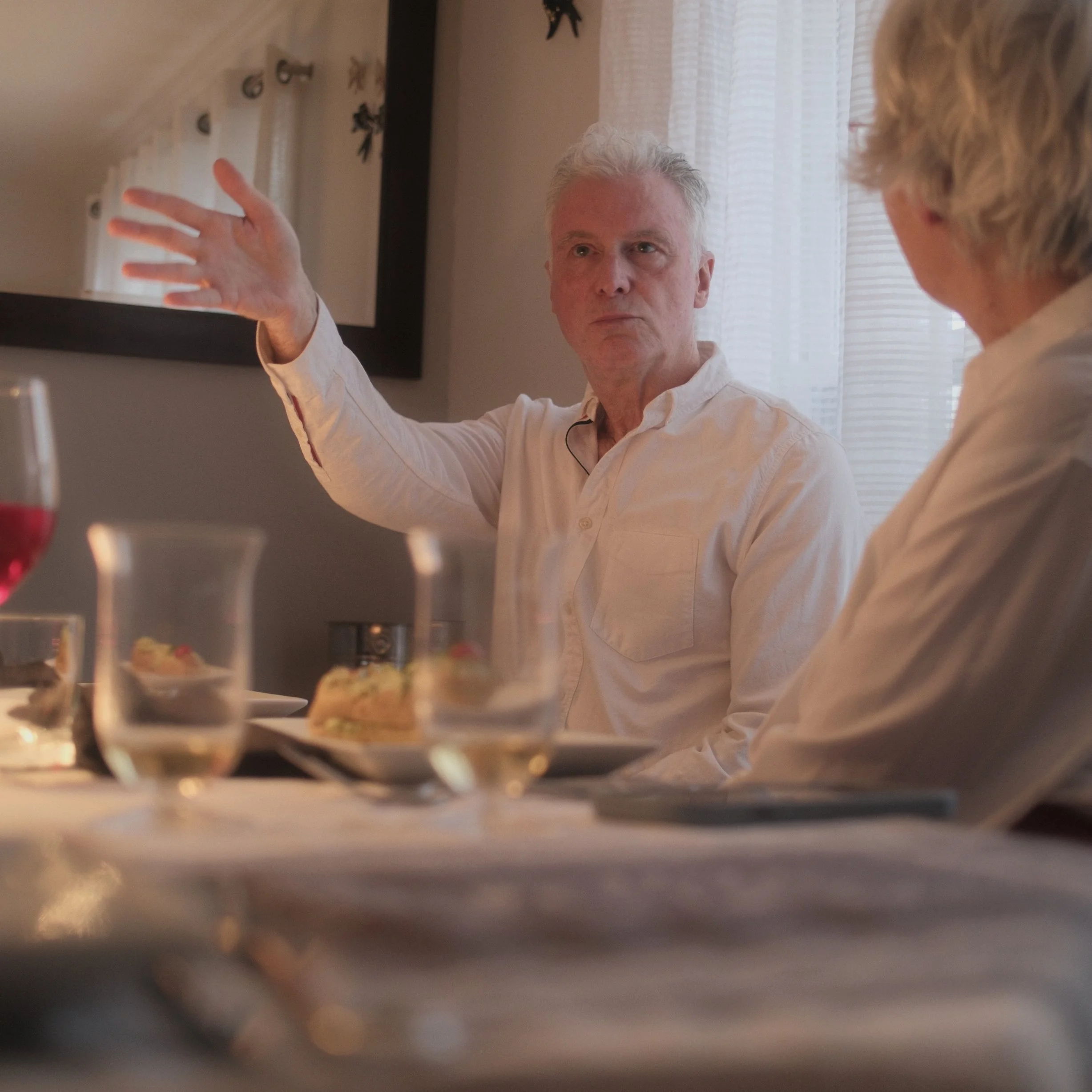 A man with gray hair gesturing with his hand during a conversation, with a woman with short blonde hair listening, in a room with curtains and wall decorations.