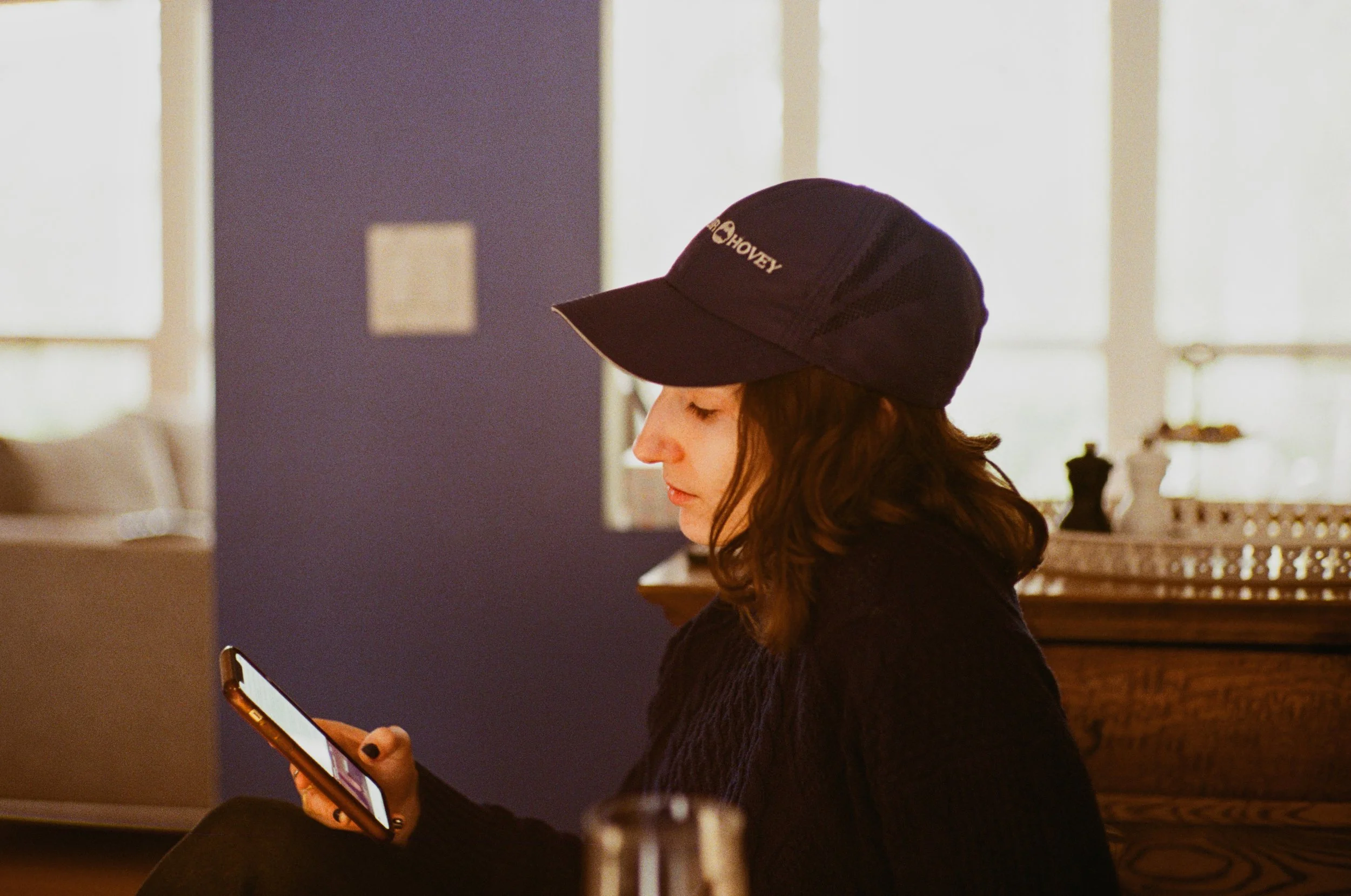 A woman with curly hair wearing a black cap with the text "Hovey" on it, looking at her phone in a cozy indoor setting.
