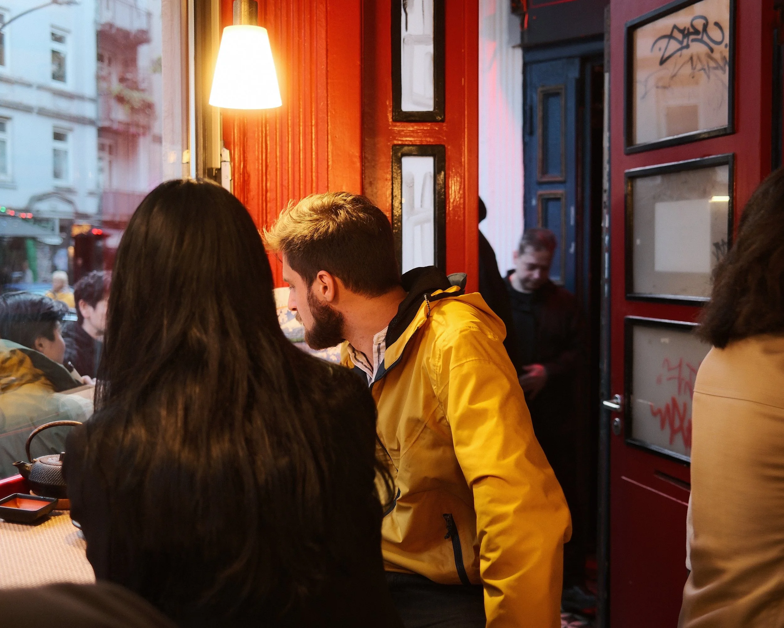 Inside a busy restaurant or cafe, several people are sitting and standing, engaging in conversations. The decor features dark frames on red walls, and warm lighting comes from a hanging lamp. Outside, a city street is visible through the windows.