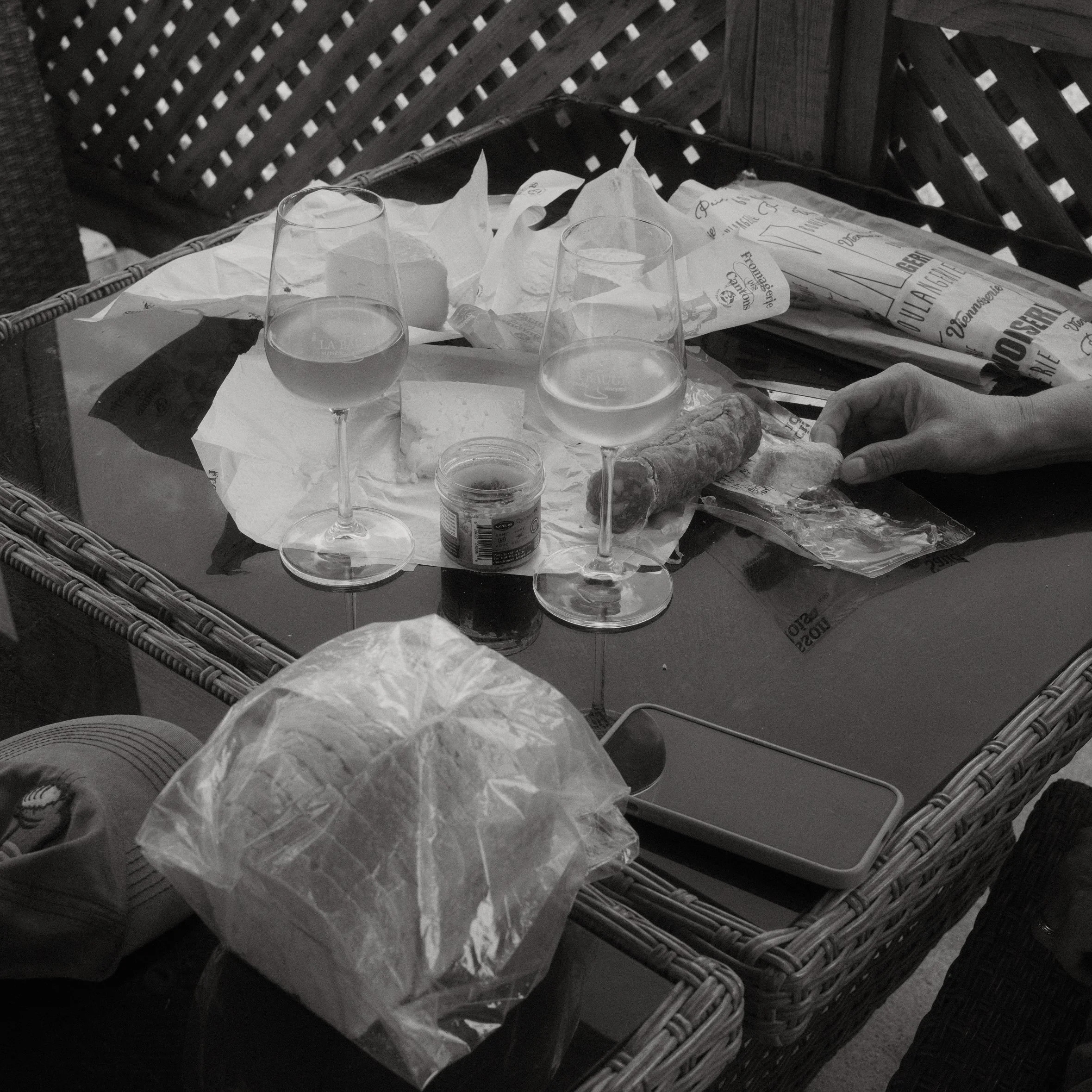 A table with three wine glasses filled with water, some crumpled paper, a newspaper, and a small jar, all placed on a tablecloth. Behind the table is a wicker chair with a wooden slatted back.