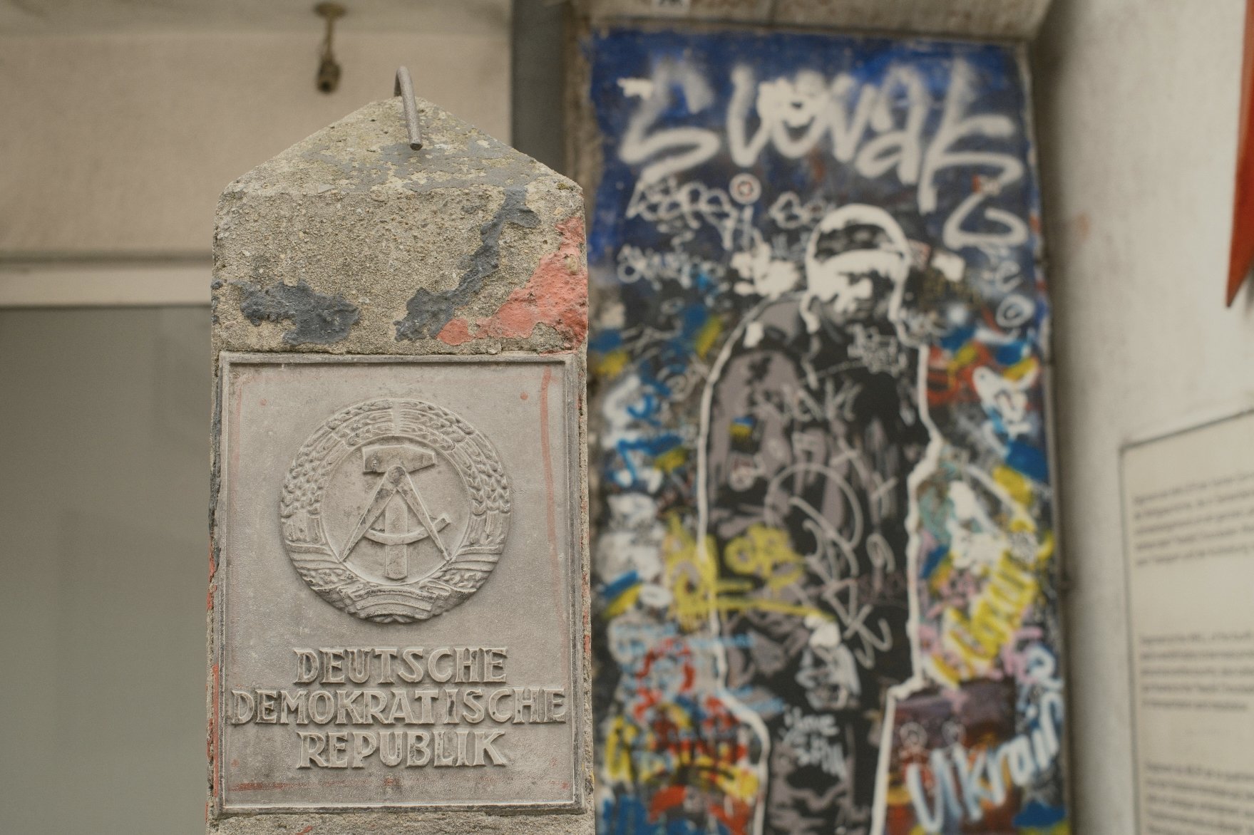 A concrete memorial plaque with the emblem of the German Democratic Republic, featuring a hammer and compass surrounded by a wreath, engraved with the words "Deutsche Demokratische Republik". In the background, a colorful graffiti-covered surface.