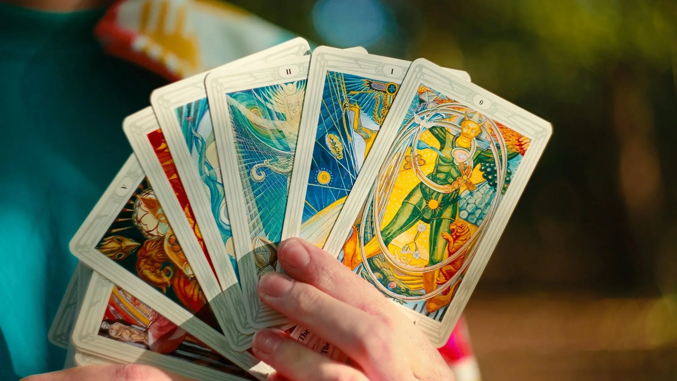 A person's hand holding a spread of colorful Thoth tarot cards with various illustrations, including a person in green, a magician, and other mystical figures and symbols.