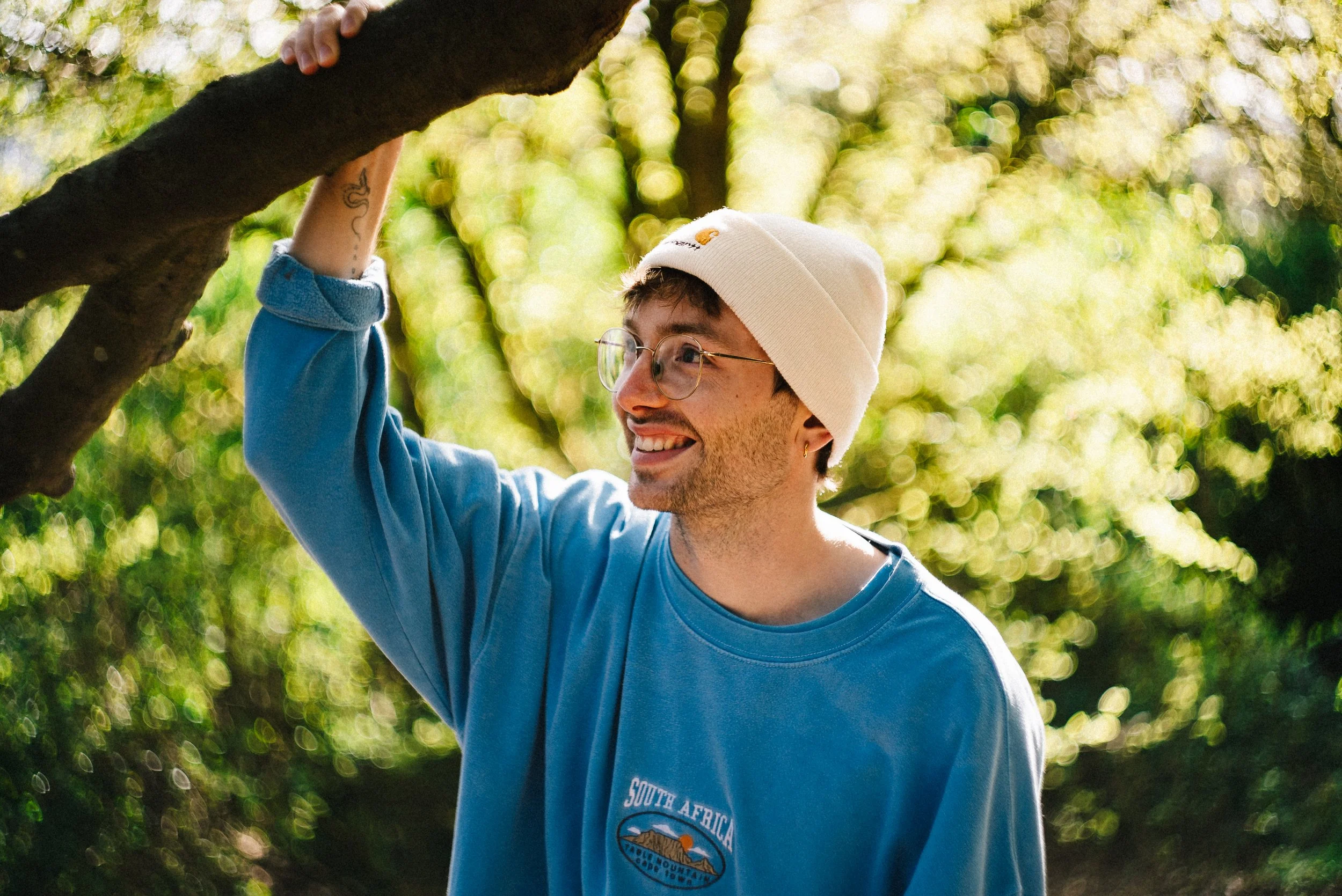 A young man with glasses and a white beanie smiling while holding a tree branch outdoors in a green, sunlit park.