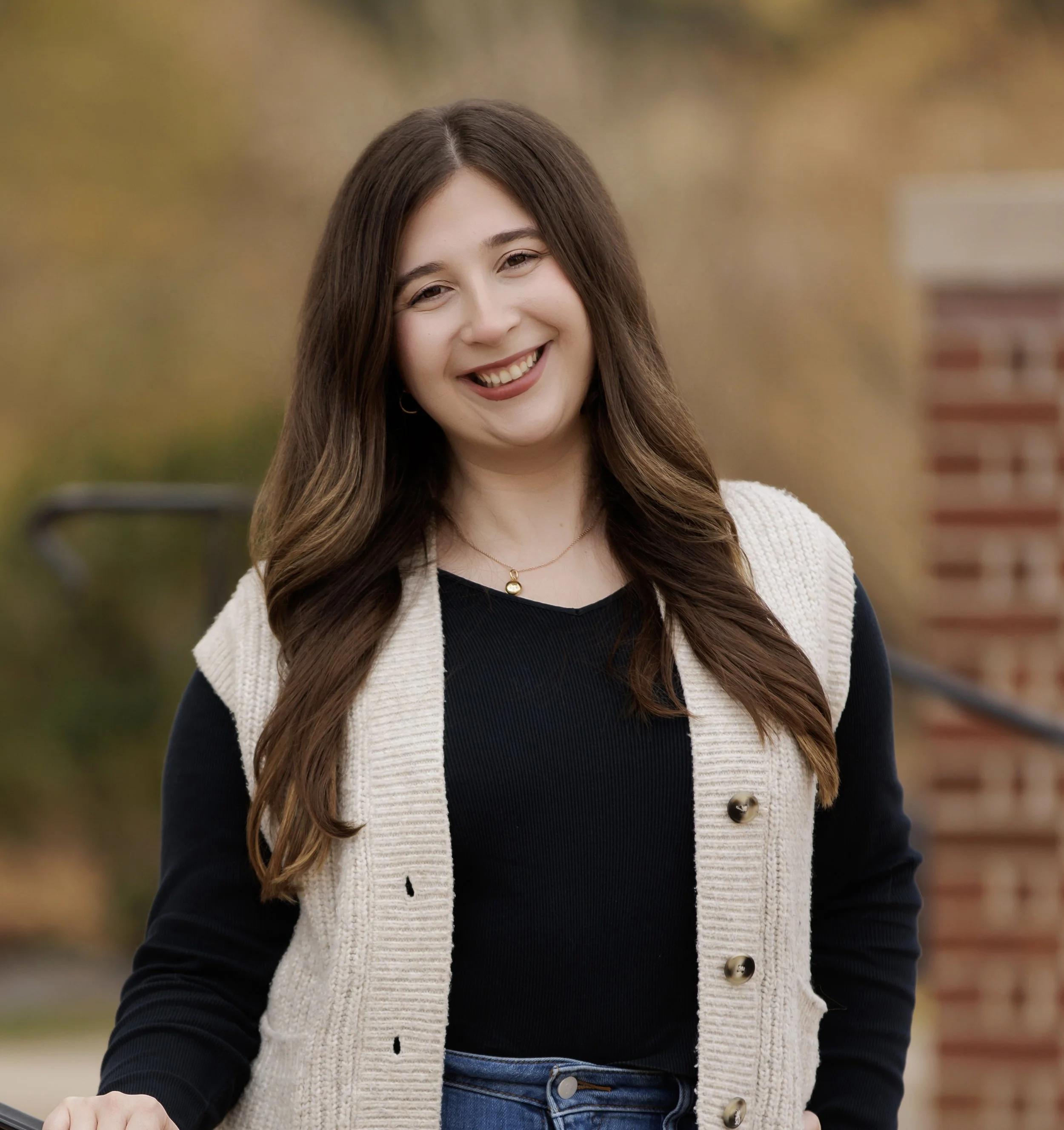 A young woman with long, wavy brown hair smiling outdoors in front of a blurred autumn background, wearing a black top, beige vest, and jeans.